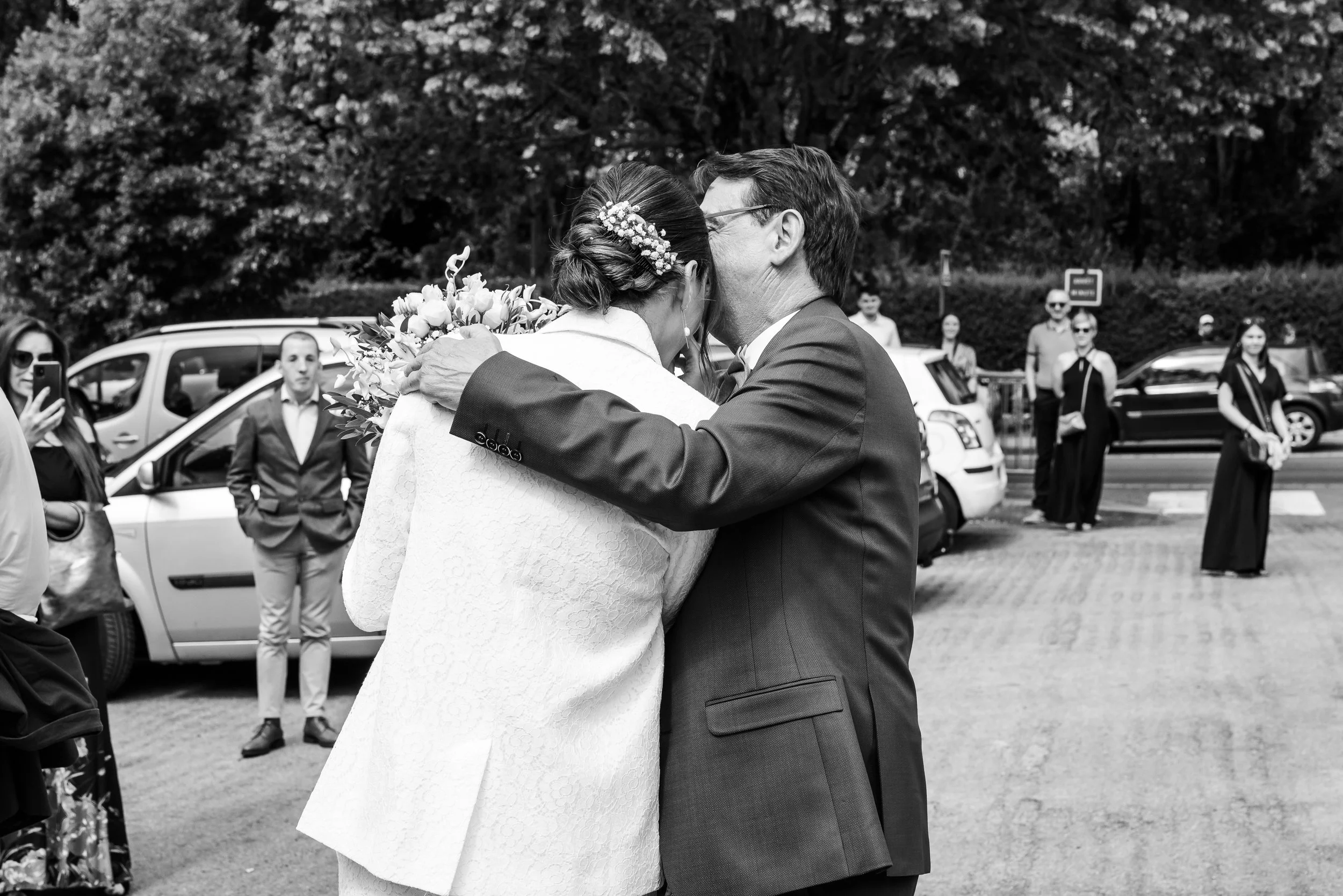 Deux personnes, un homme et une femme, s'embrassent à une fête ou un mariage. La femme porte une robe blanche et une coiffure ornée de fleurs, et tient un bouquet de fleurs. L'homme porte un costume sombre. Plusieurs personnes en arrière-plan regarde