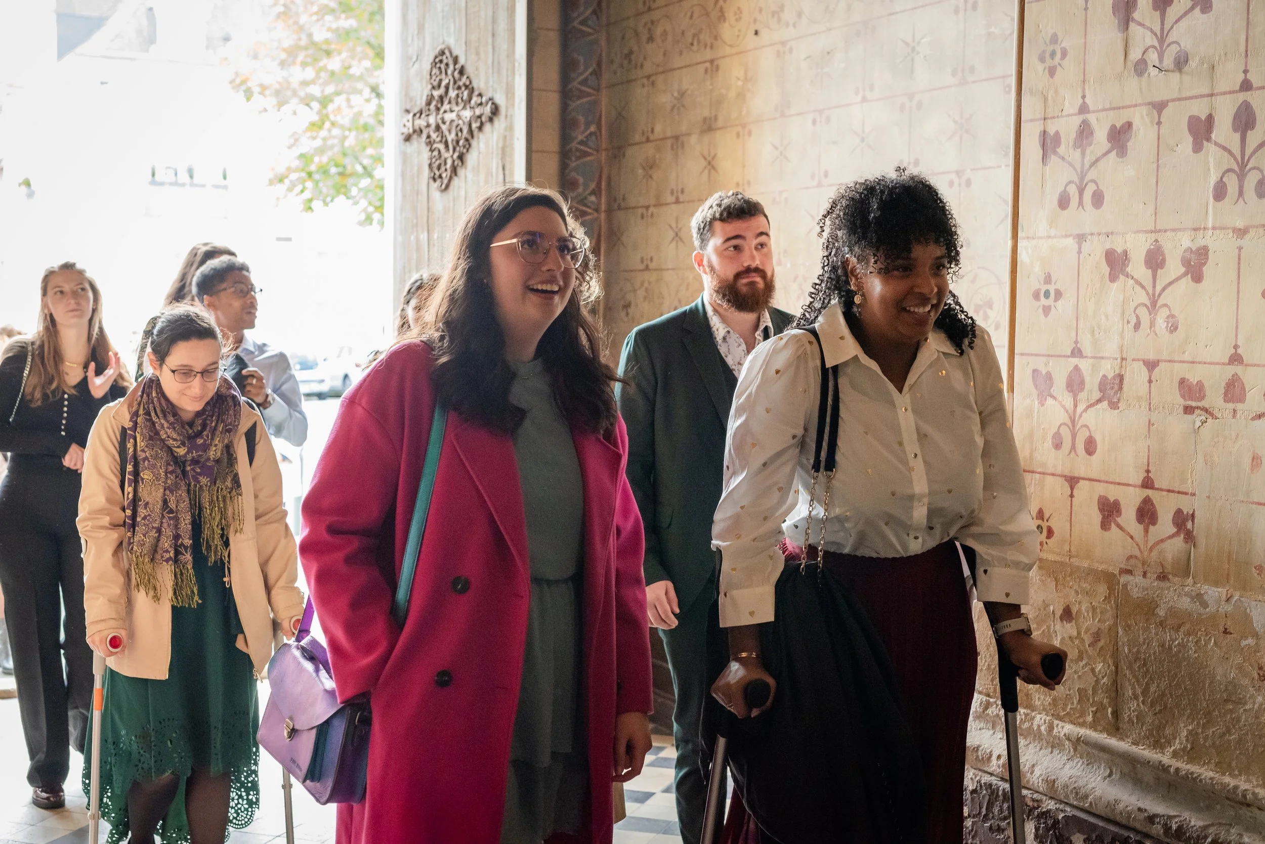 Groupe de personnes entrant dans un bâtiment historique avec décoration ancienne, certaines souriantes, une femme utilise une canne et porte un sac.