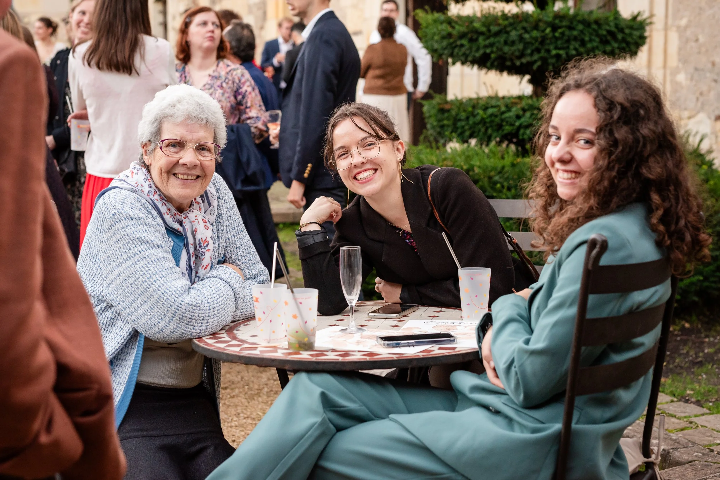 Trois femmes souriantes assises autour d'une table lors d'une réunion en extérieur, avec des invités en arrière-plan, dans un jardin ou une cour pavée.