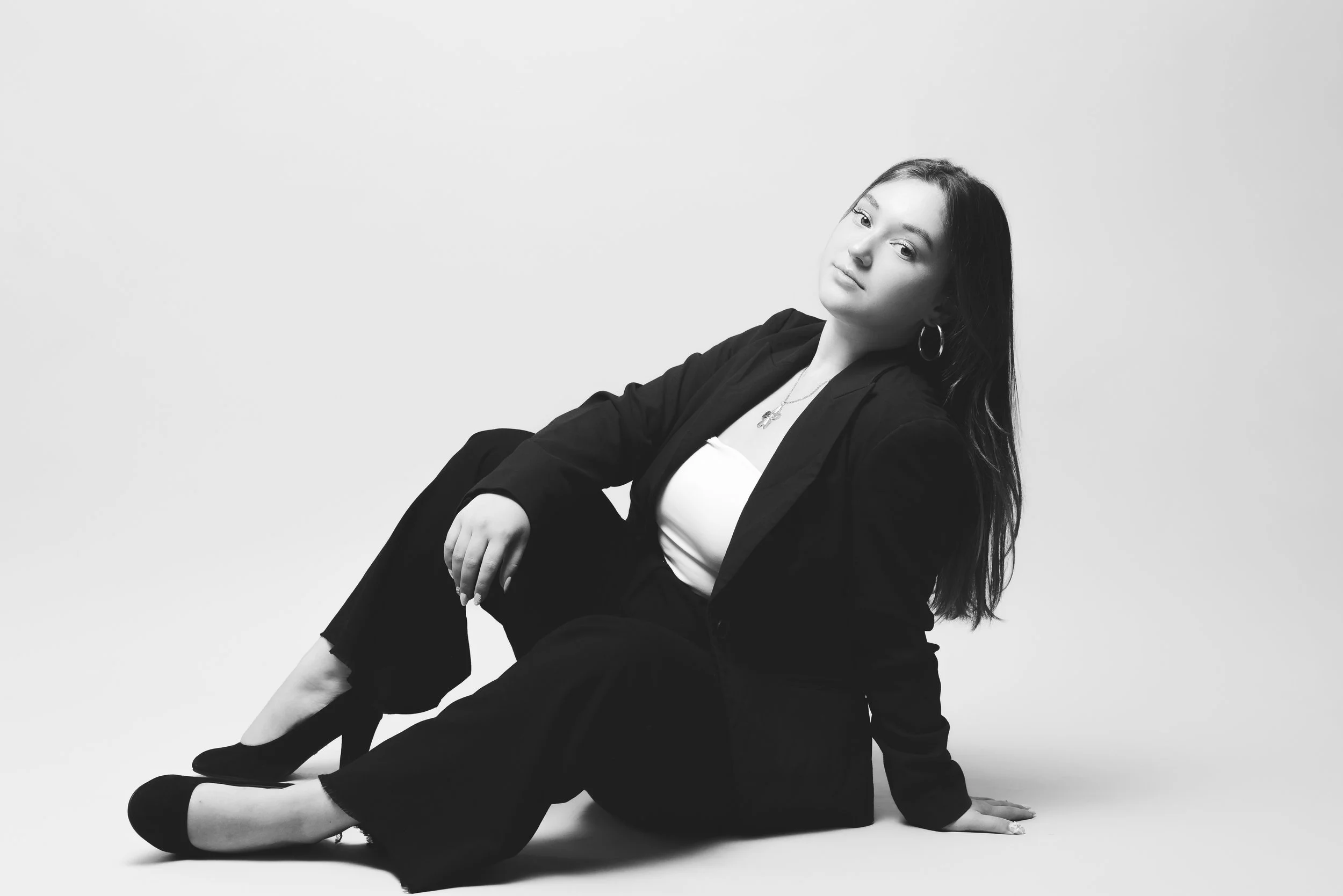 Femme assise au sol en noir et blanc, portant un blazer noir, une robe blanche, des boucles d'oreilles, et un collier, posant contre un mur blanc.