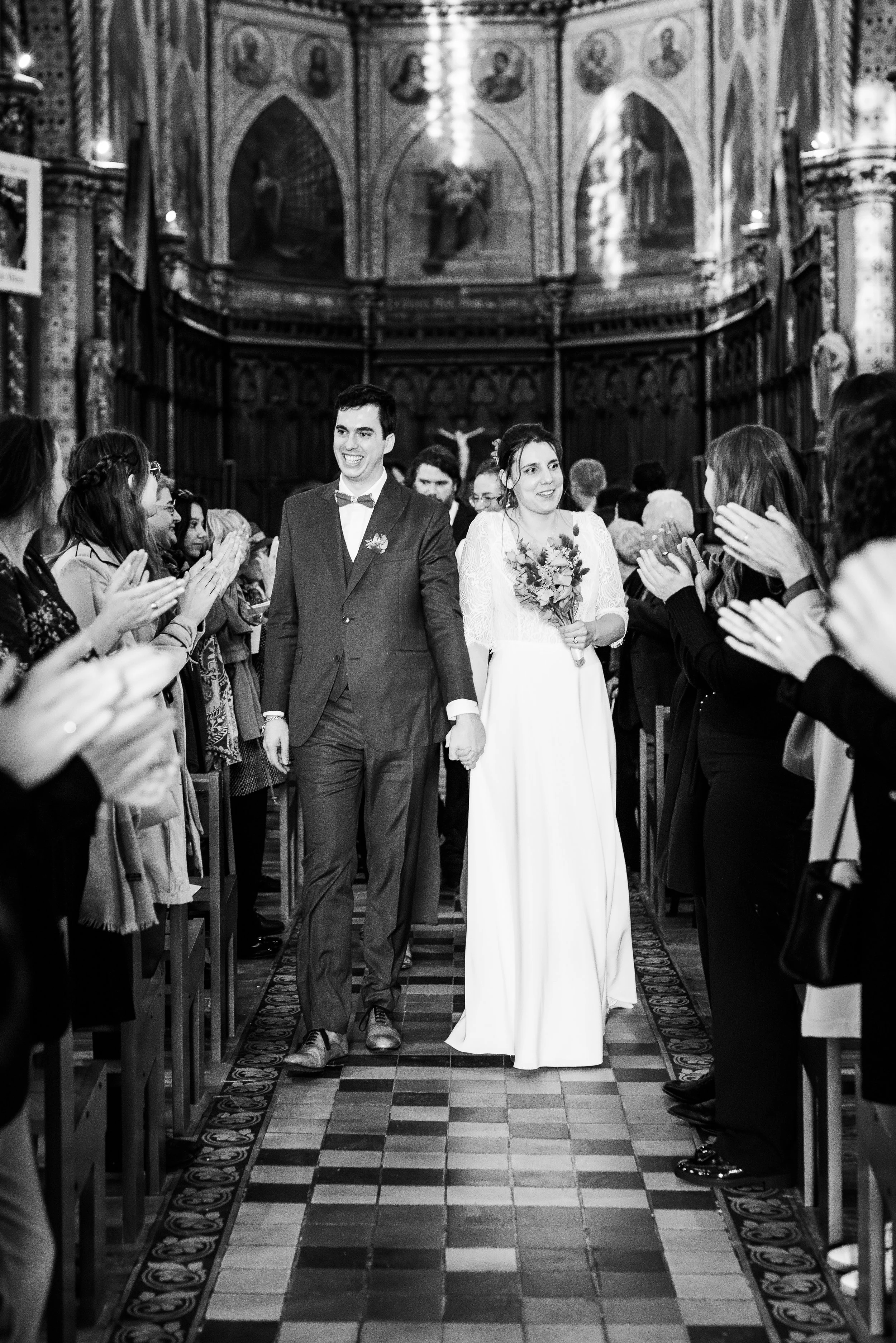 Un mariage dans une église, couple marchant main dans la main au centre, entouré d'invités applaudissant, décor religieux en arrière-plan.