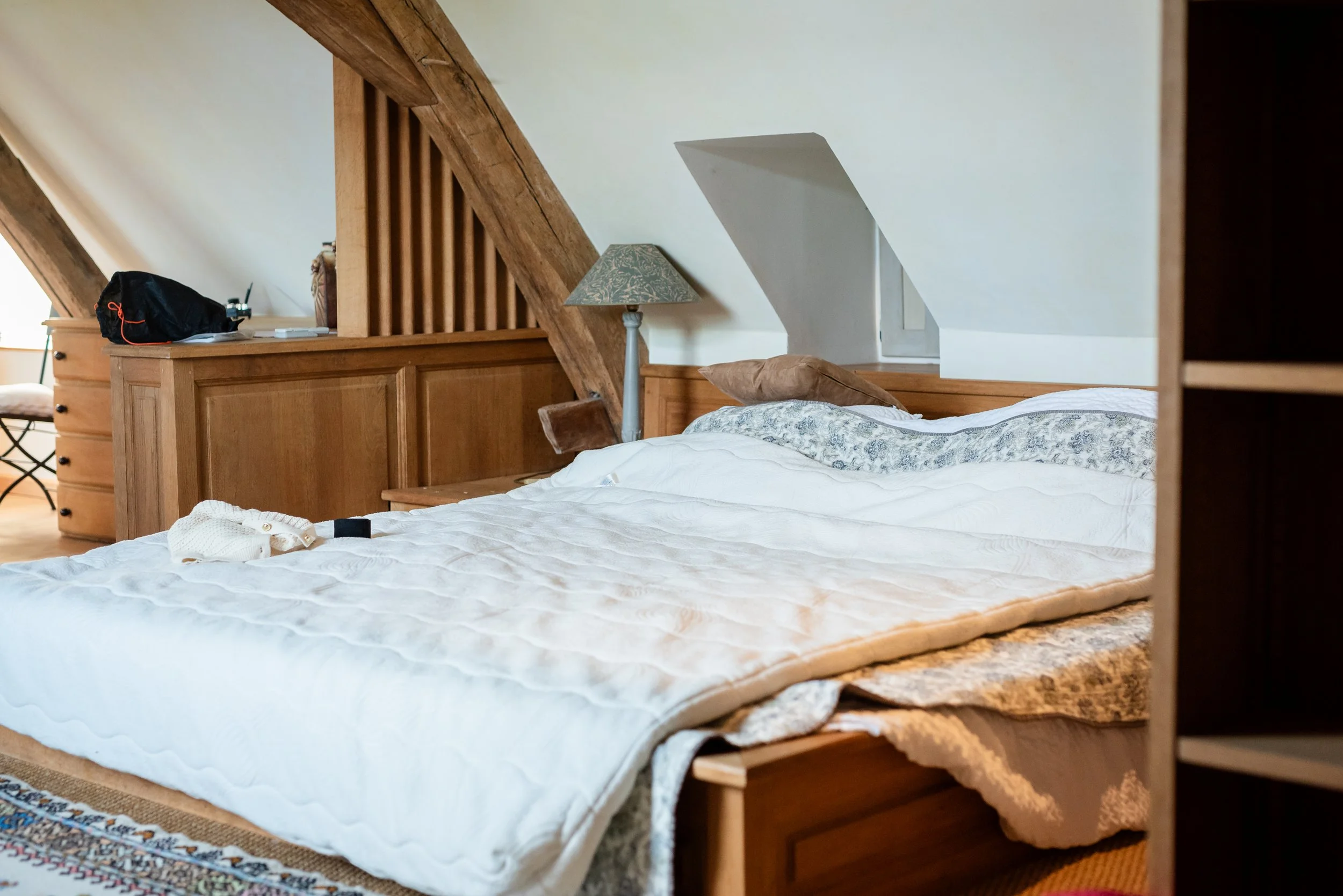 Lit simple avec un oreiller beige, dans une chambre mansardée en bois avec un lampadaire et des meubles en bois.