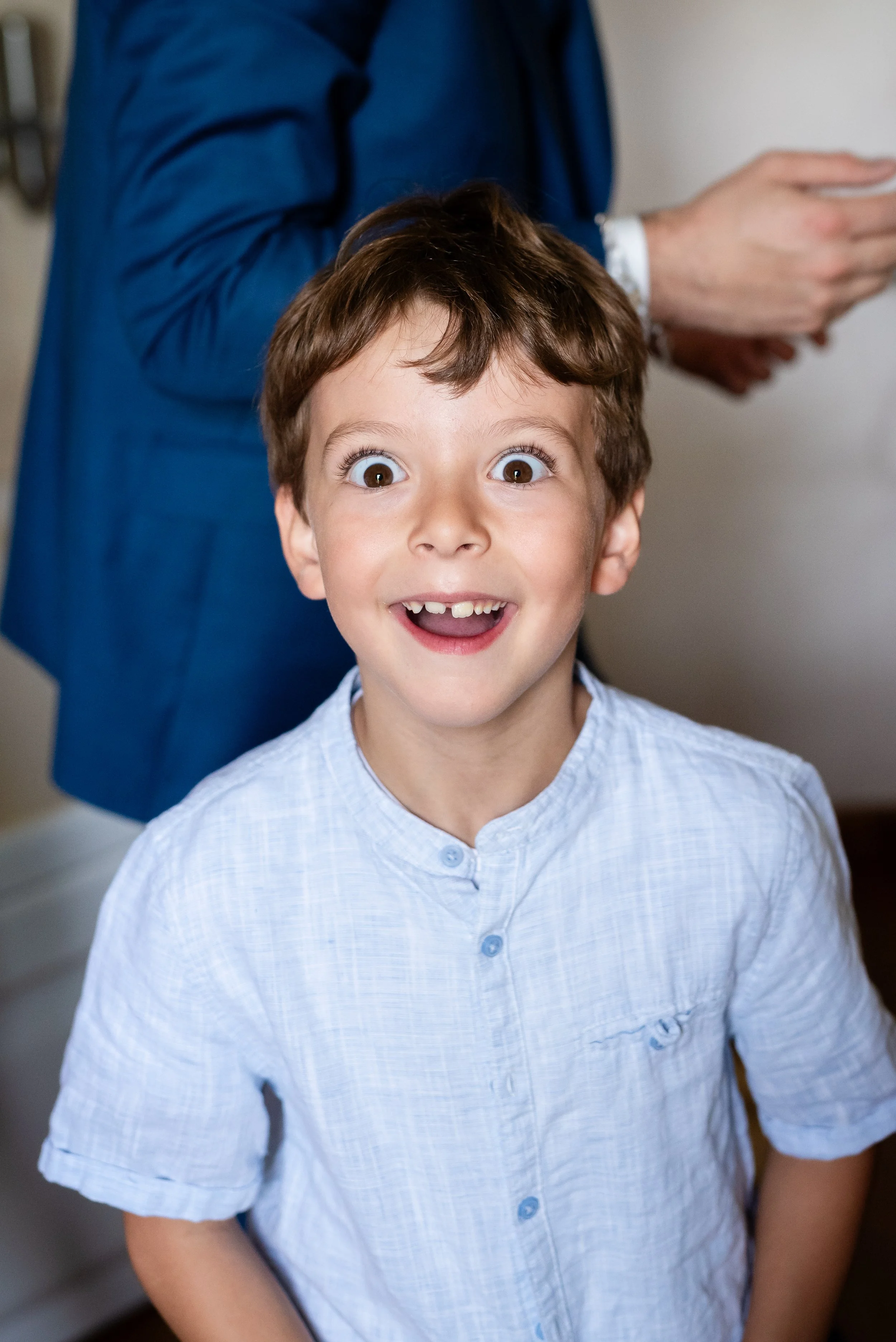 Un garçon souriant avec des yeux grands et une dent manquante, portant une chemise blanche à manches courtes. Un adulte, vraisemblablement un parent, en arrière-plan pousse un smartphone.