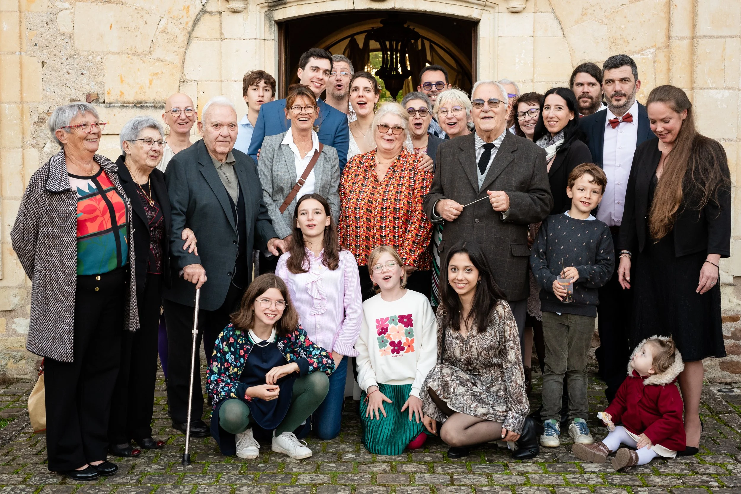 Un groupe de personnes de différentes générations posant devant une vieille façade en pierre, certains souriant, d'autres en train de parler ou faisant des expressions variées.