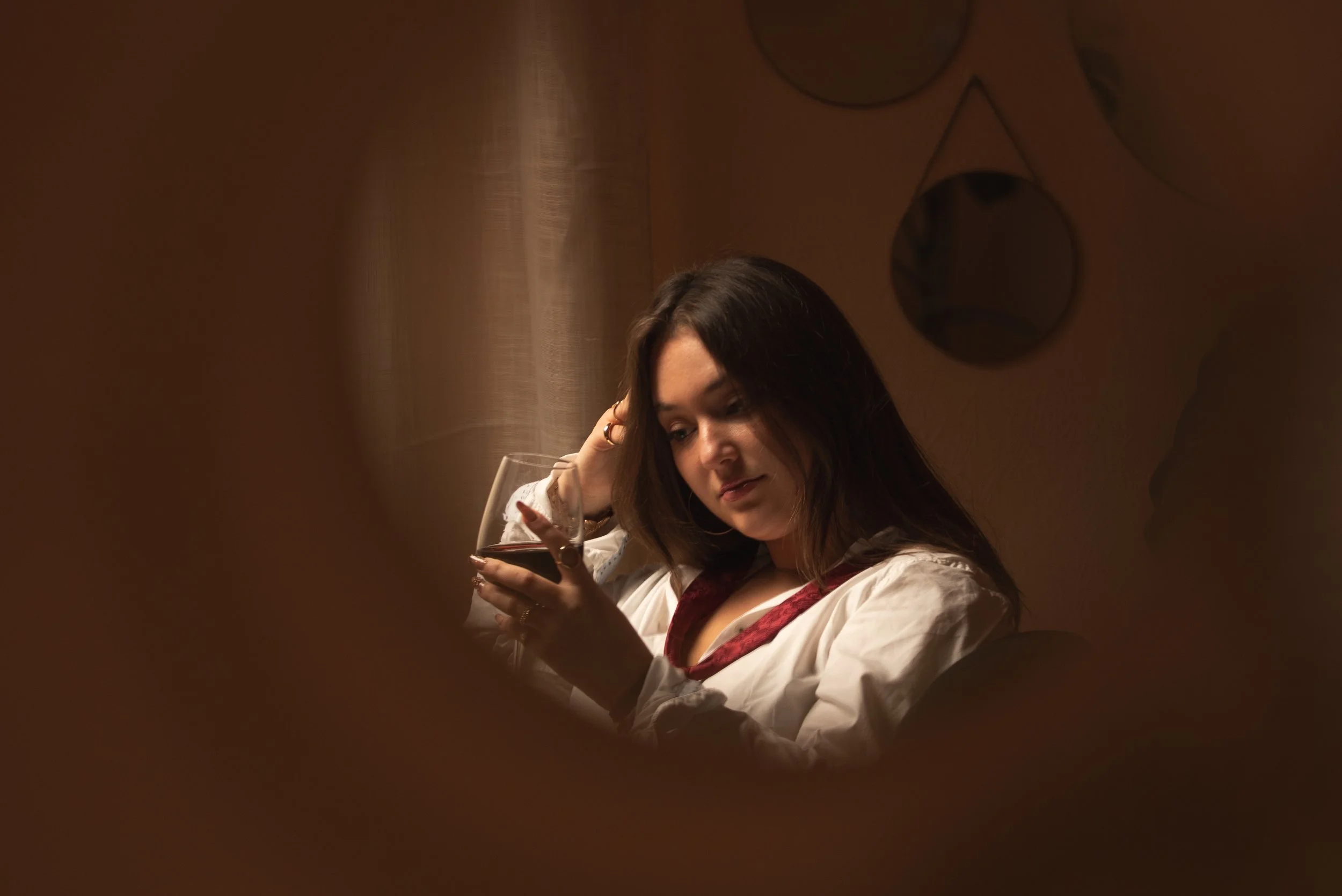 Une femme avec des cheveux bruns longs, portant une veste blanche avec des détails rouges, regarde un verre de vin rouge qu'elle tient dans sa main, dans un environnement intérieur avec un mur beige et des décorations murales rondes.