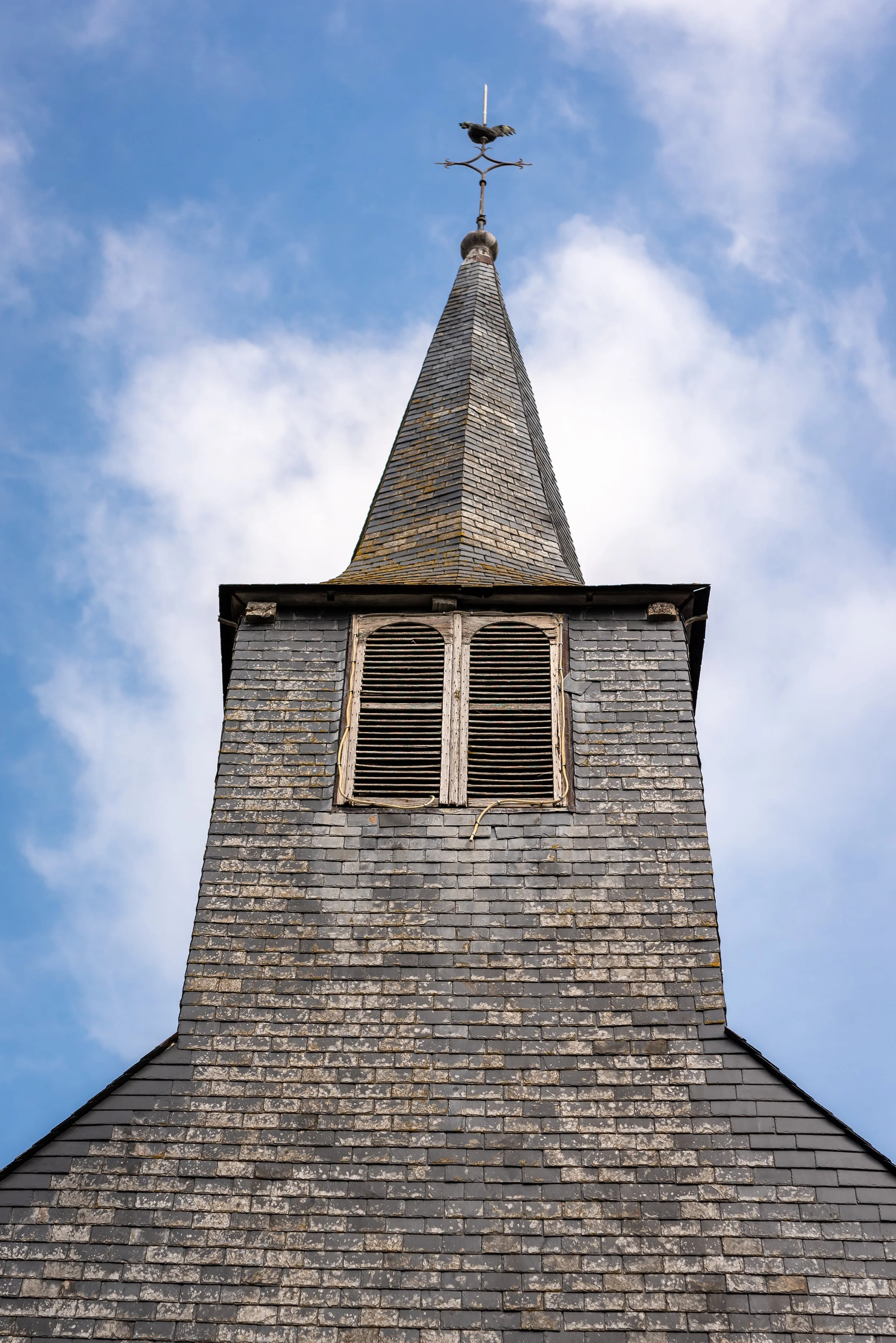 Mât de clocher en ardoise avec une girouette en forme de corbeau au sommet, contre un ciel bleu avec quelques nuages.