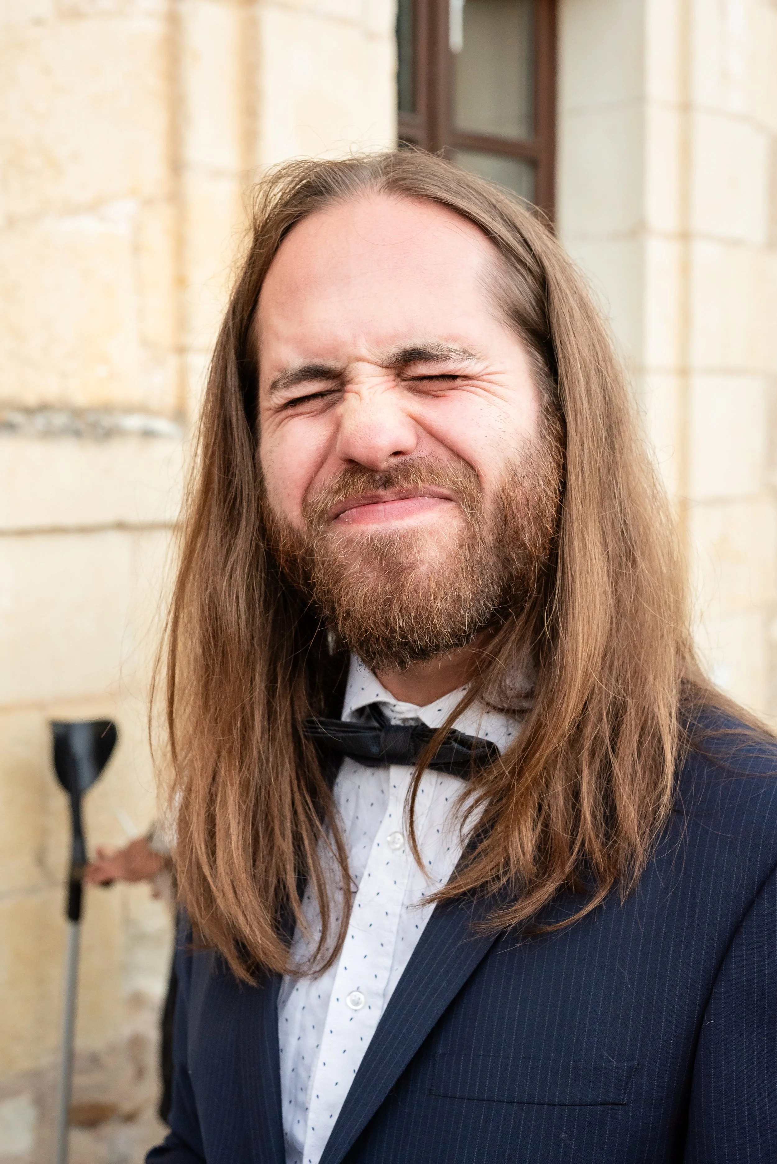 Un homme avec de longs cheveux et une barbe, souriant avec les yeux fermés, portant un costume à rayures et une chemise à pois, en arrière-plan un mur jaune et une fenêtre.