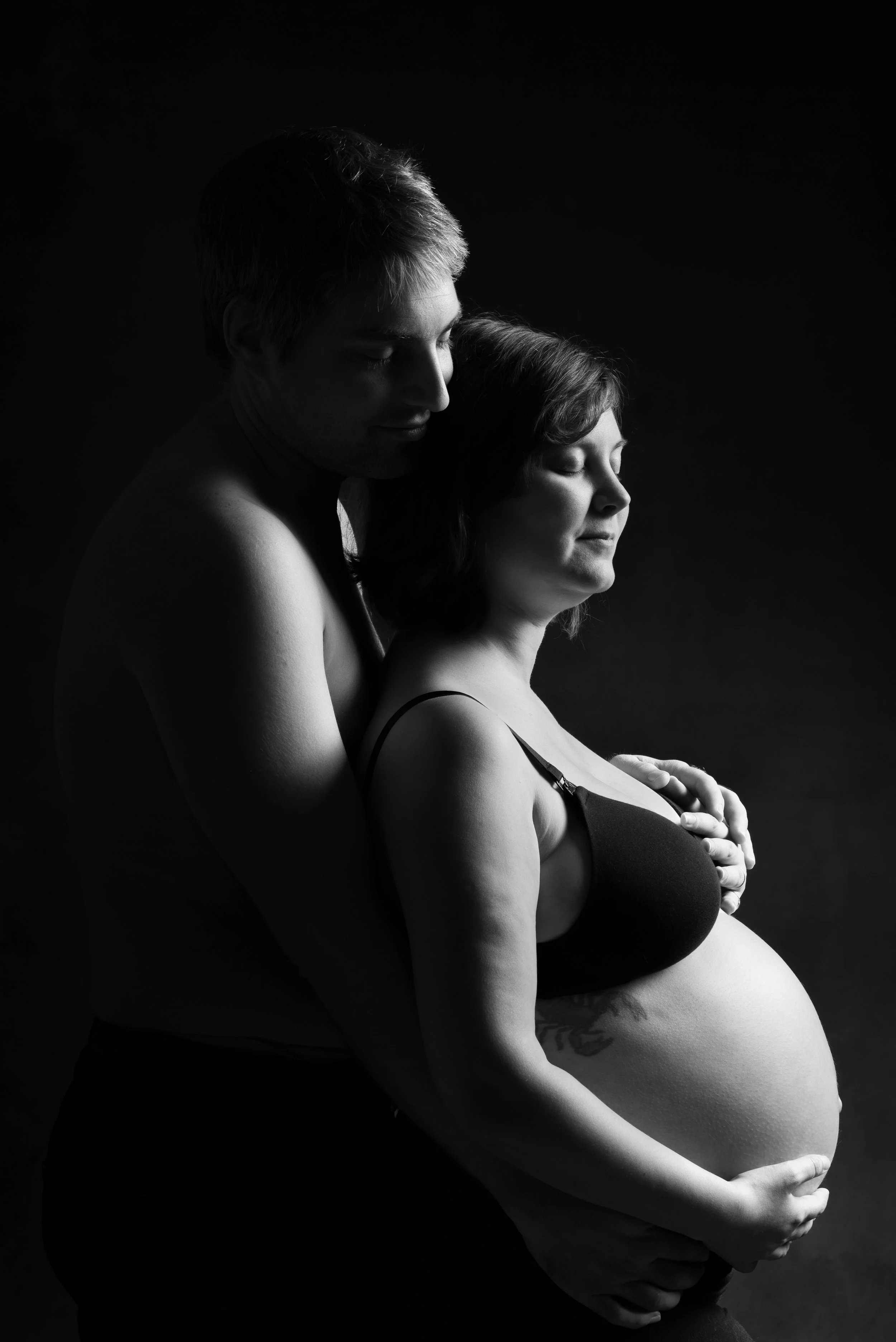 Une femme enceinte habillée en noir et un homme nue, dans une pose tendre avec des yeux fermés, en noir et blanc.