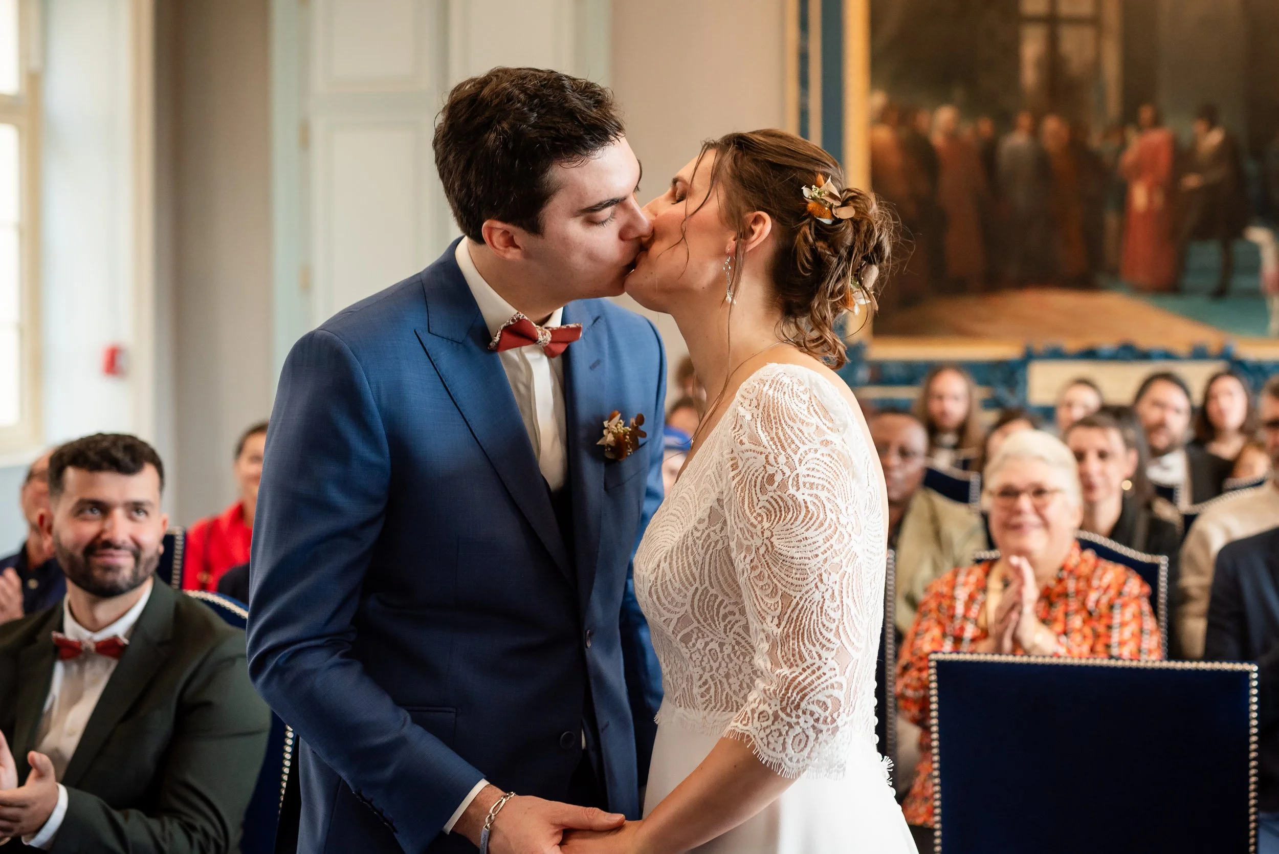 Un couple en mariage s'embrassant lors de leur cérémonie, assis dans une salle avec des invités en arrière-plan.