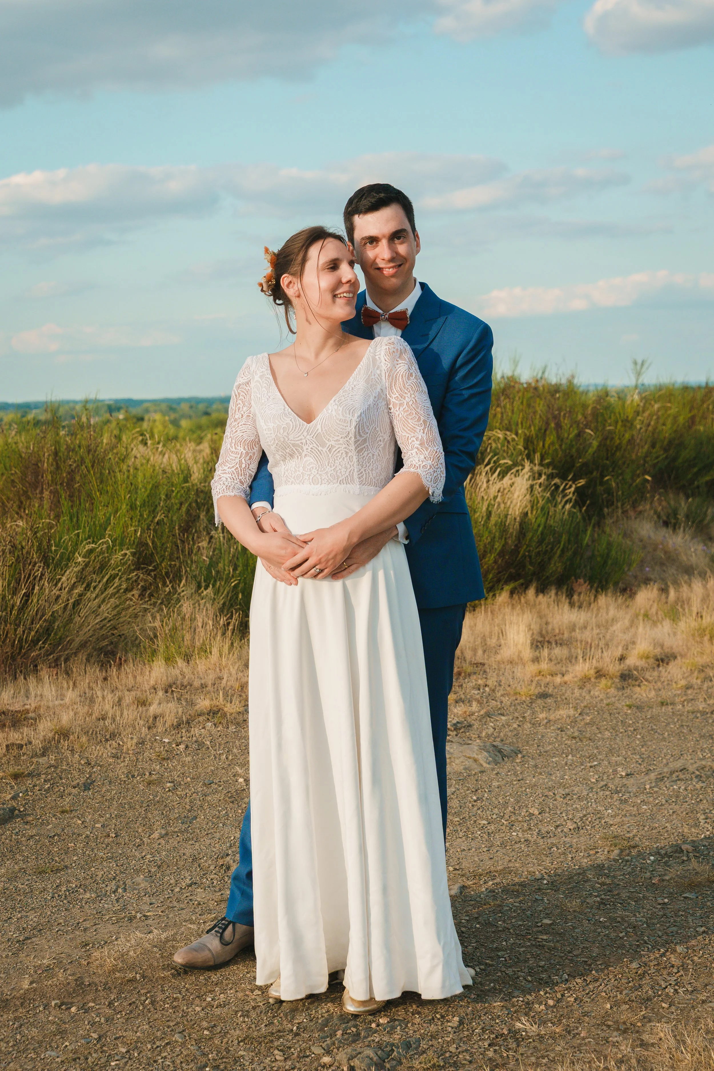 Un couple marié en plein air, la femme sourit et regarde à gauche, l'homme regarde droit devant, derrière un ciel bleu avec quelques nuages, dans un champ avec des herbes hautes.