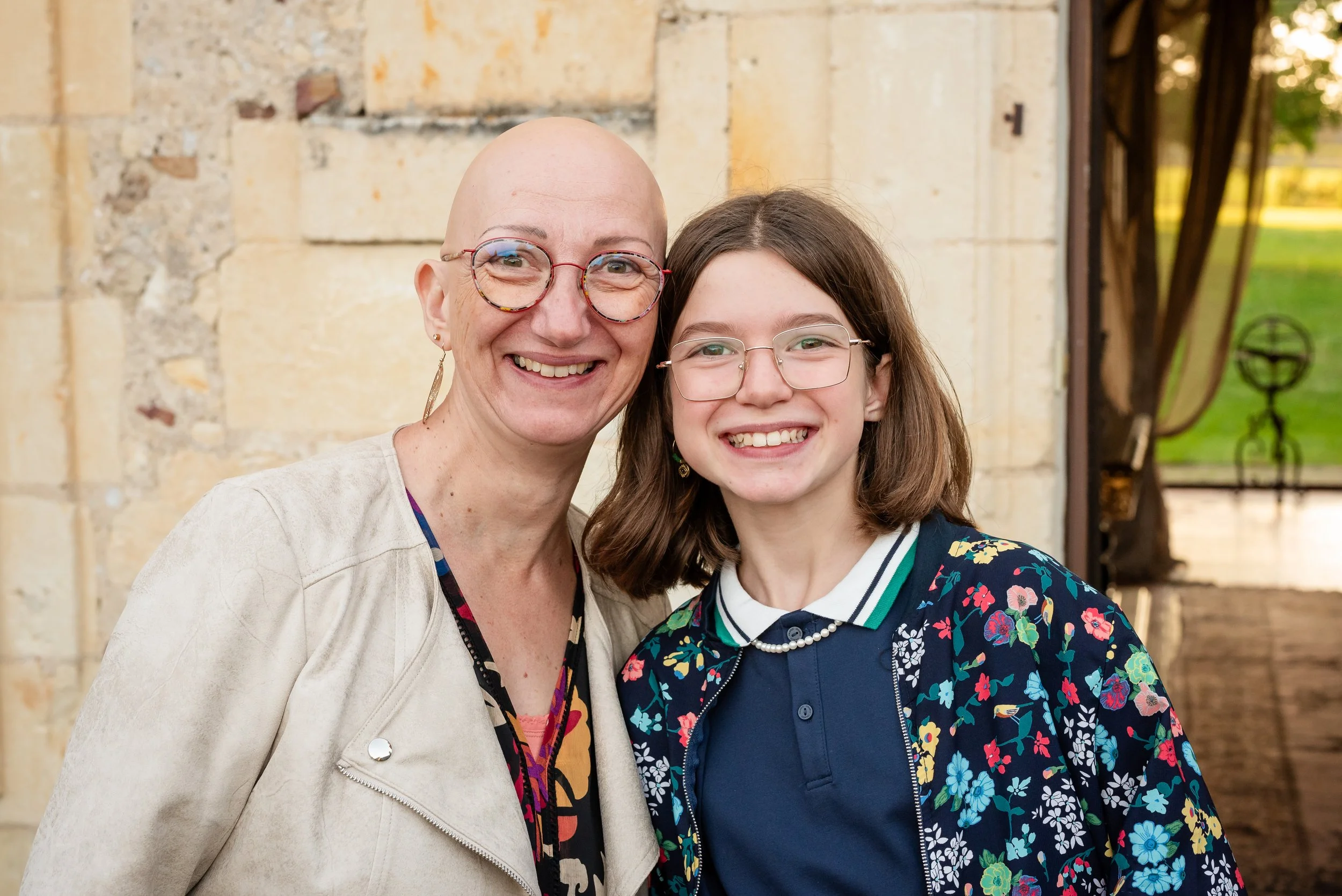 Deux femmes souriantes posent ensemble devant un bâtiment en pierre, une porte en bois visible à l'arrière.