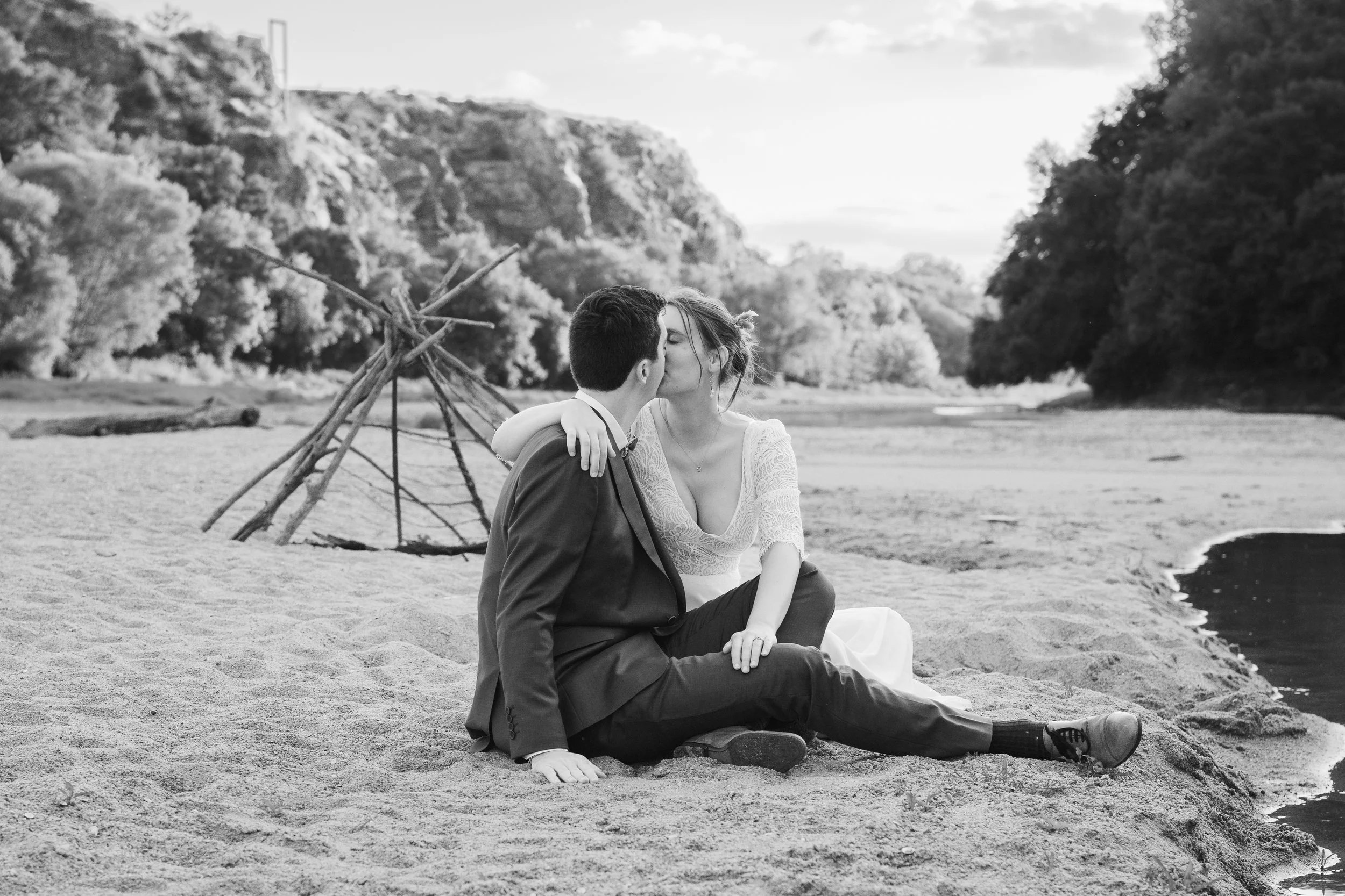 Un couple assis sur la plage, se donnant un baiser, avec un décor naturel comprenant des arbres et un rivage d'eau en arrière-plan, en noir et blanc.