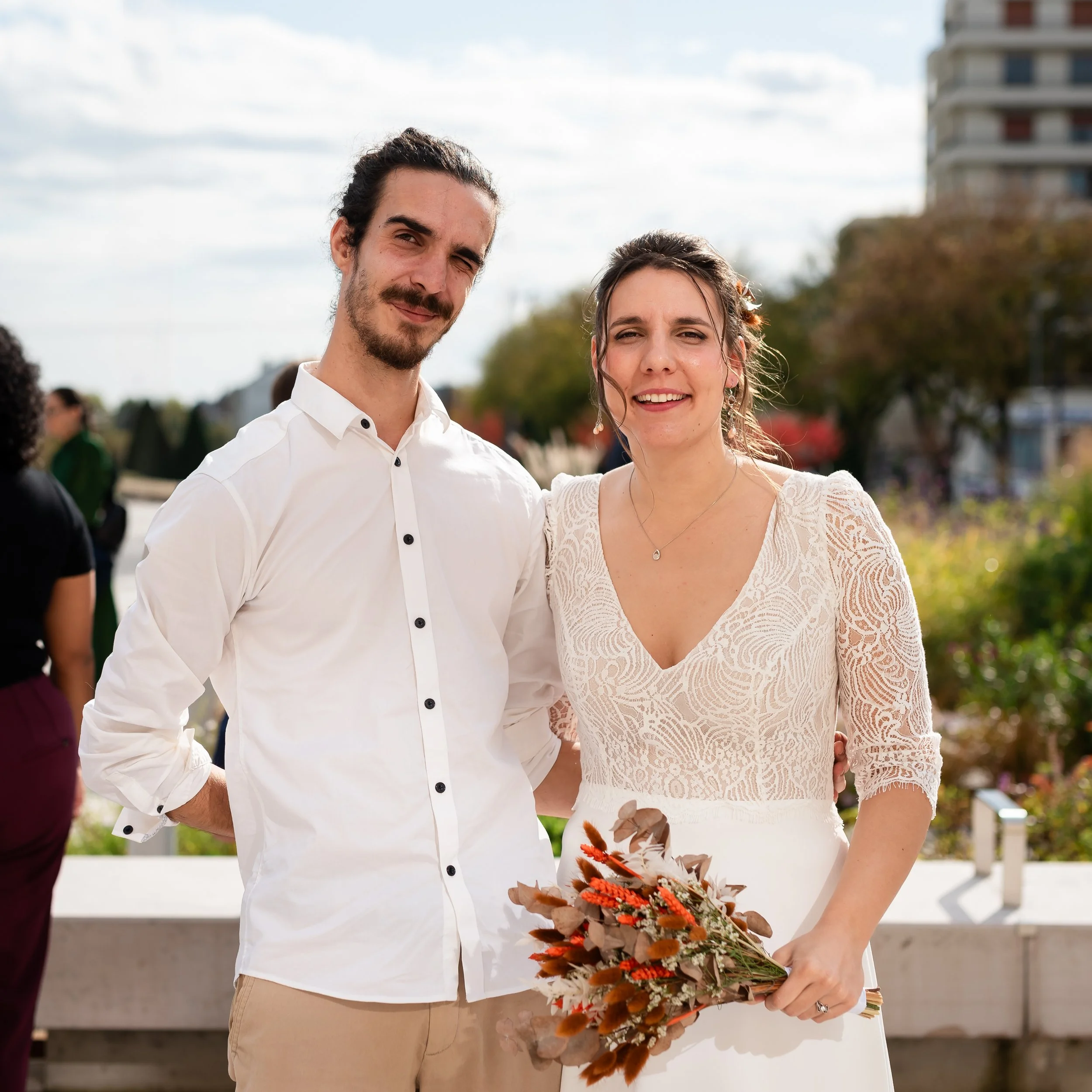 Un homme et une femme debout ensemble, tenant un bouquet de fleurs sèches, lors d'une cérémonie, en plein air, par une journée ensoleillée.