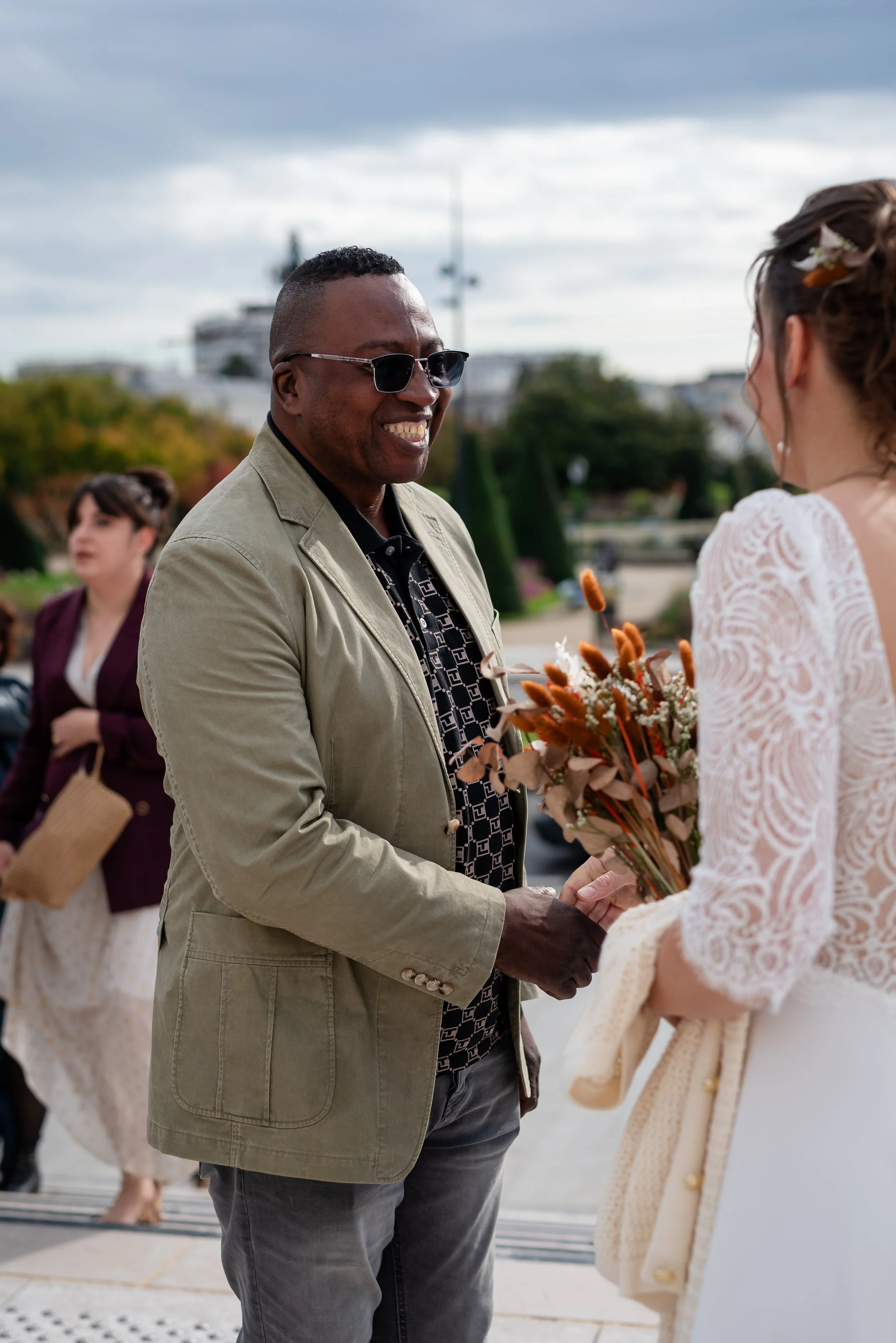 Un homme souriant portant des lunettes de soleil, en train de remettre un bouquet de fleurs à une femme lors d'une cérémonie en plein air.