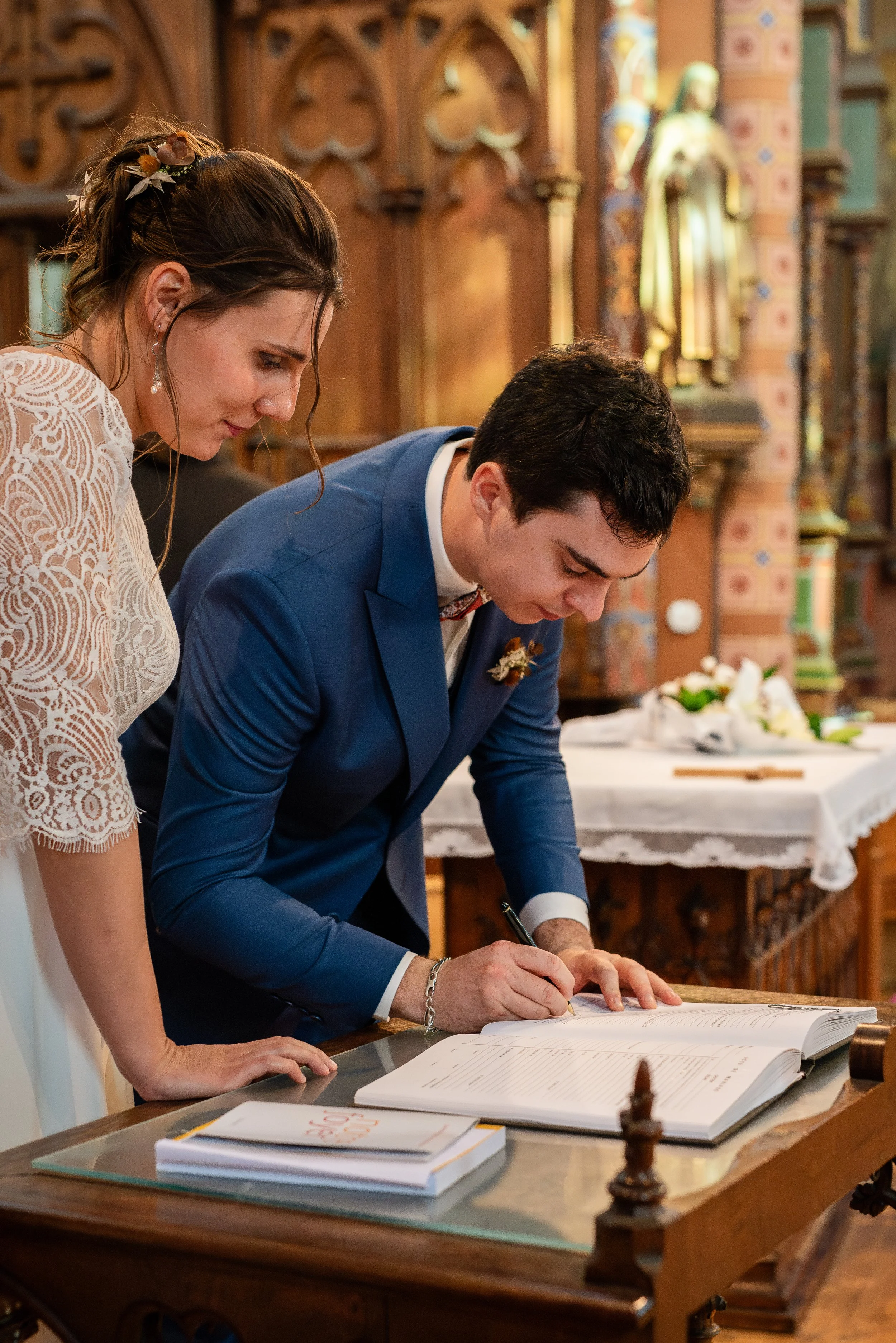 Un homme en costume bleu écrit dans un livre, tandis qu'une femme en robe blanche en dentelle regarde. La scène semble se dérouler dans une église ou une salle décorée avec des éléments religieux.