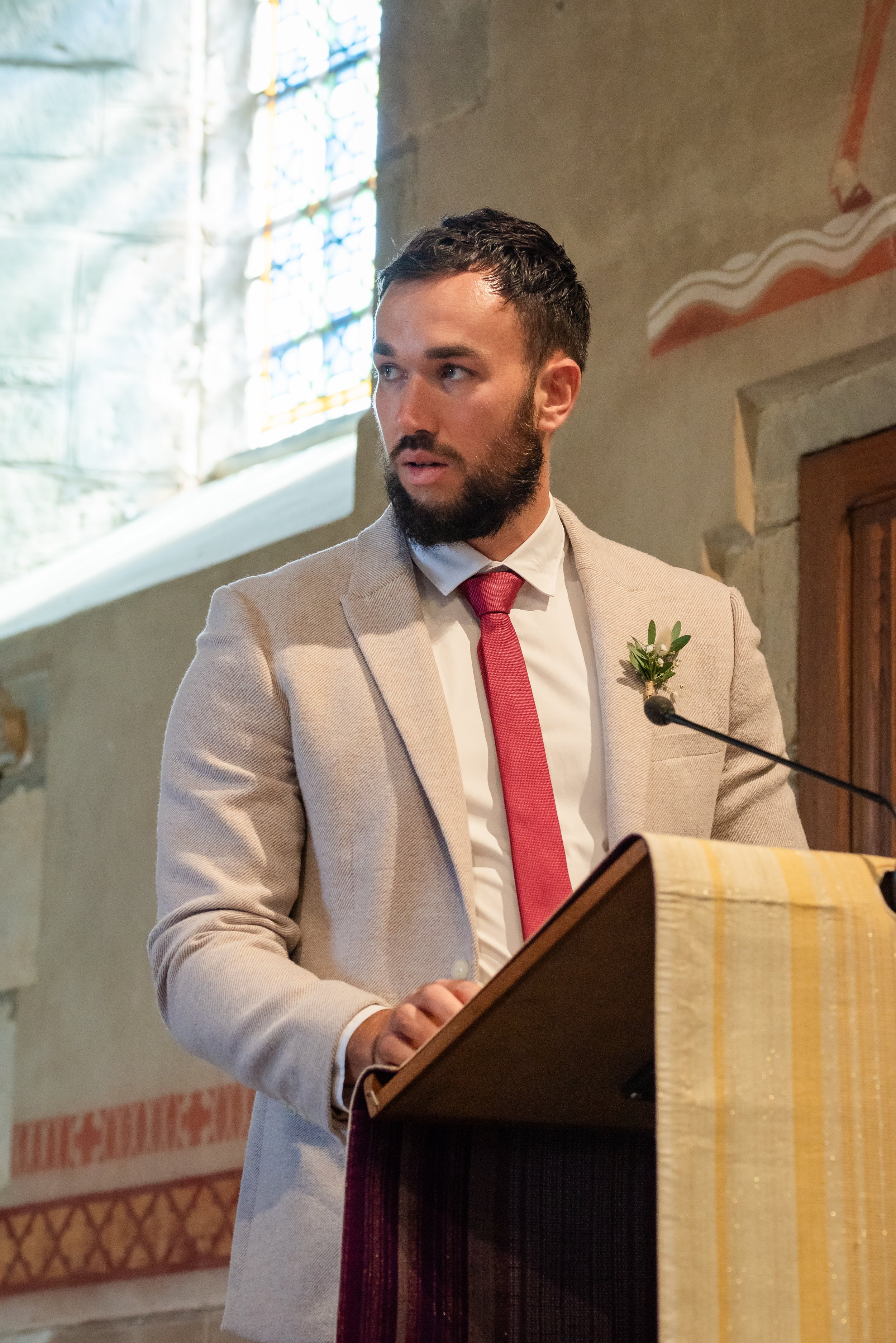 Un homme avec une barbe, portant un costume beige, une chemise blanche et une cravate rouge, est en train de parler ou de faire un discours dans une église.