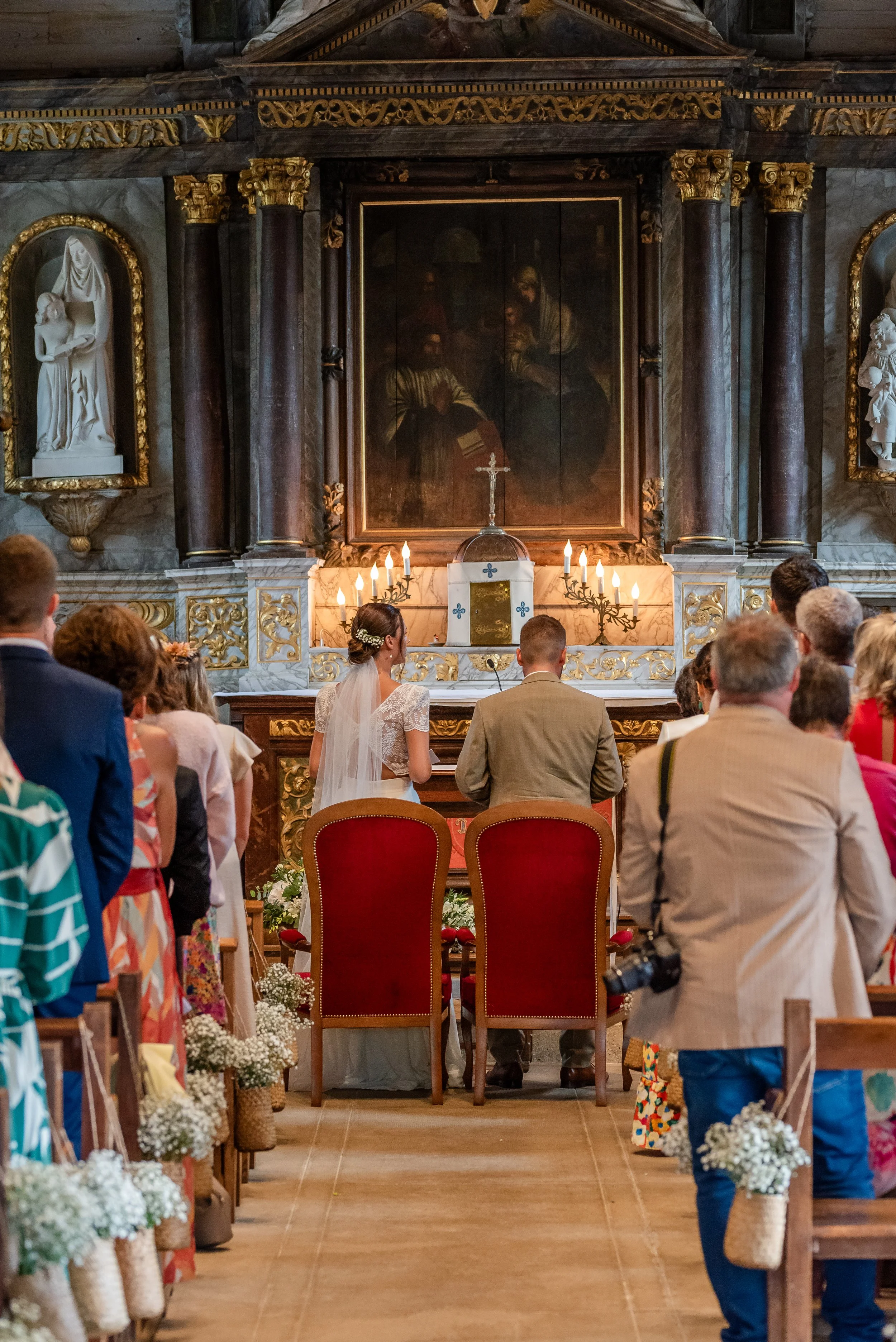 Cérémonie de mariage dans une église avec des invités assis, une mariée et un marié au premier plan, des décorations florales, un autel et des crucifix en arrière-plan.
