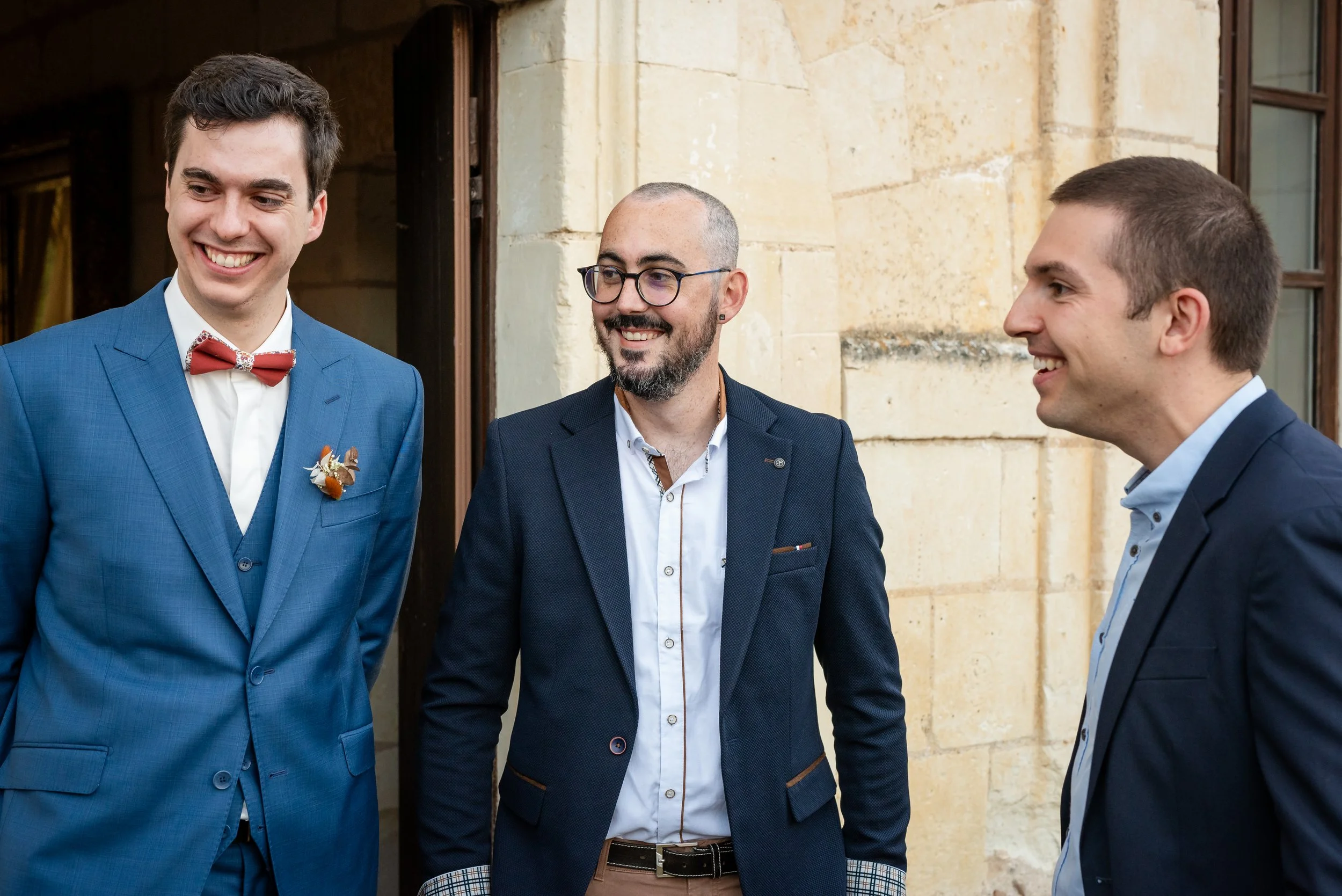 Trois hommes habillés en costumes sourient et discutent devant un bâtiment en pierre.