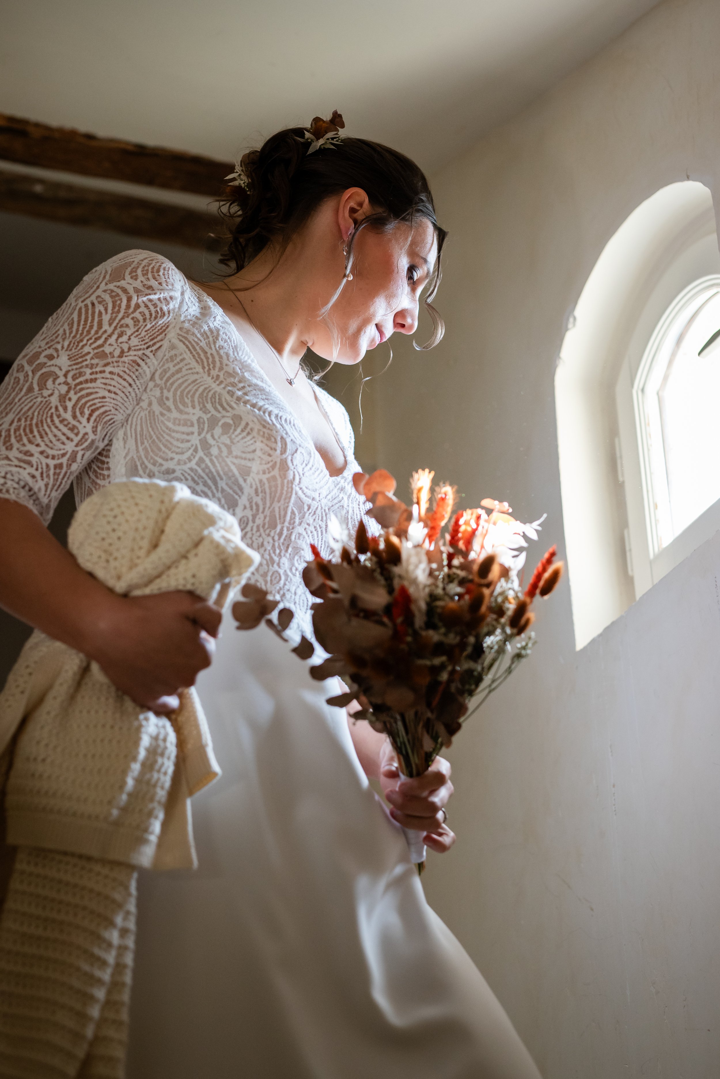 Jeune femme habillée en robe de mariage, tenant un bouquet de fleurs séchées, devant une fenêtre dans une pièce lumineuse.