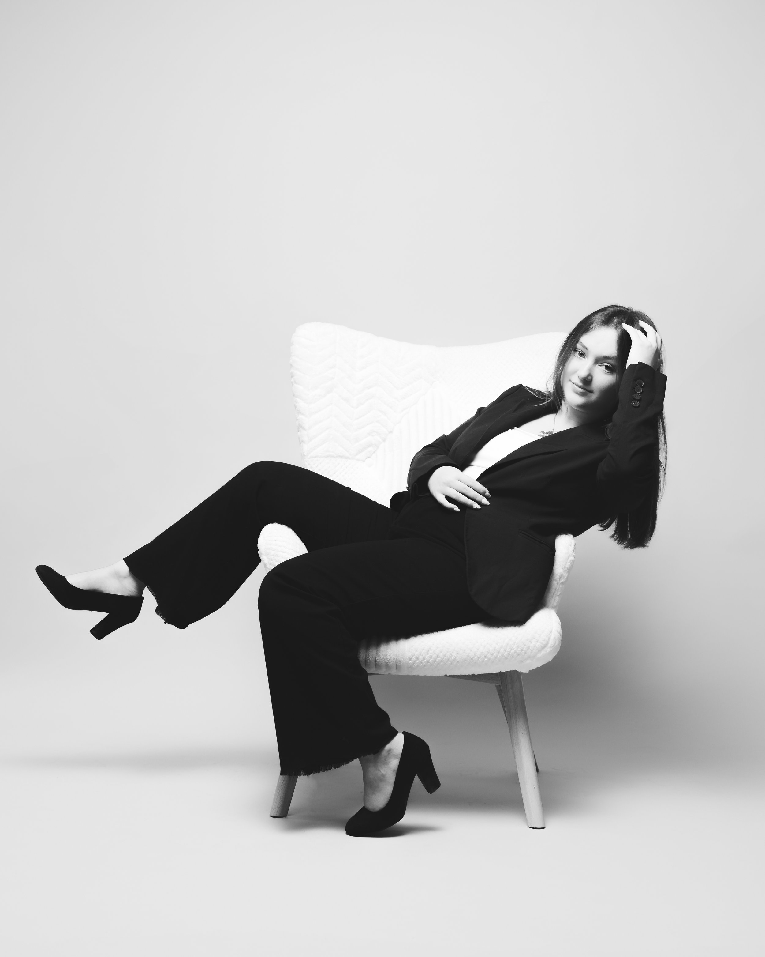 Une femme assise sur une chaise blanche, portant un tailleur noir, avec ses jambes croisées et des talons noirs, dans un studio avec un fond blanc.