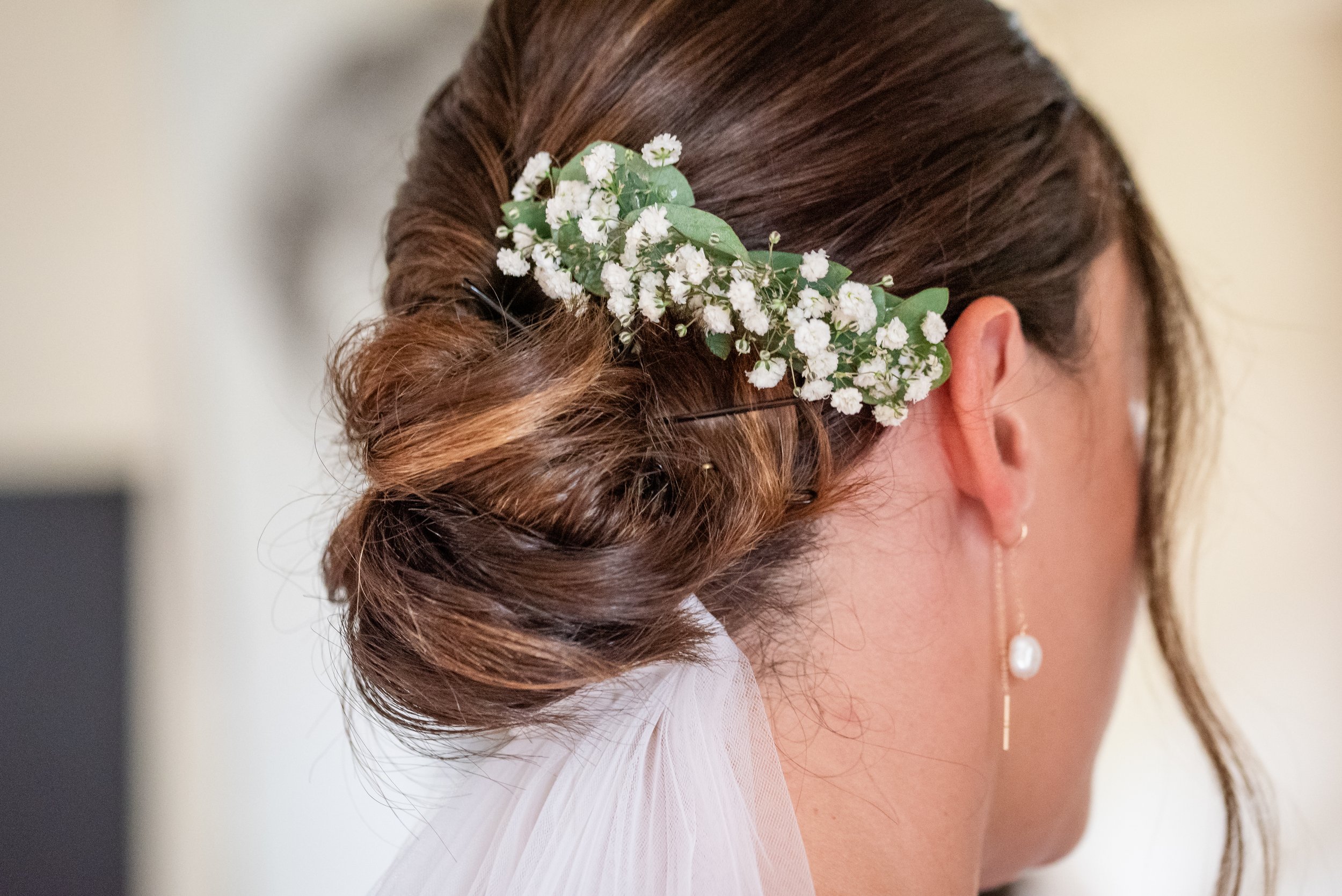 Coiffure de mariage avec des cheveux châtains coiffés en un chignon bas orné d'une petite branche de fleurs blanches et de feuilles vertes, avec une boucle d'oreille pendante en perle visible.