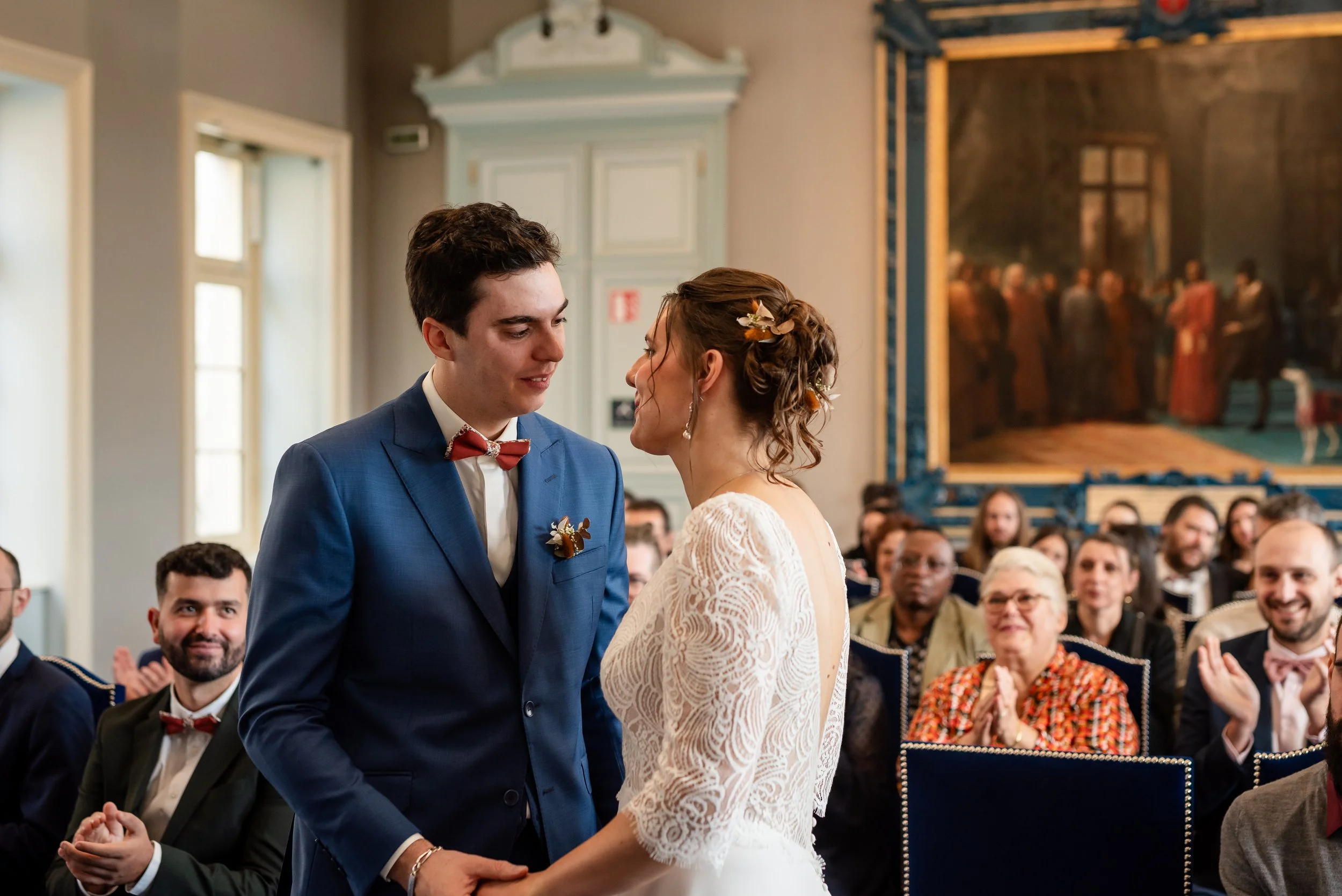 Un couple lors de leur mariage échangeant un regard lors de la cérémonie, avec des invités qui observent en arrière-plan dans une salle élégante.