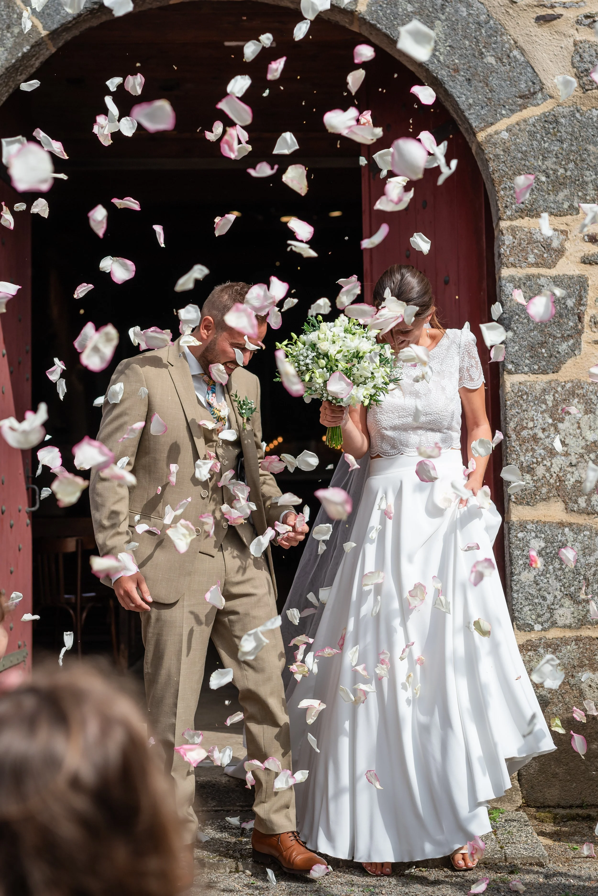 Un couple en mariage sortant d'une porte en pierre, entouré de pétales de fleurs tombés.