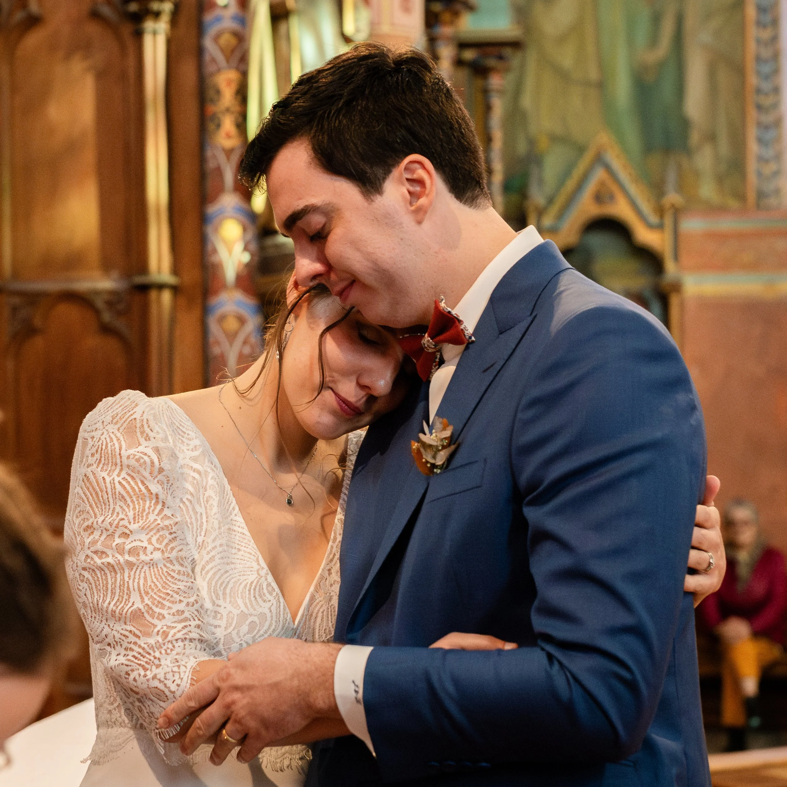 Un couple se tient très proche lors d'une danse, dans un endroit décoré avec des motifs colorés. La femme porte une robe en dentelle blanche et des lunettes, tandis que l'homme porte un costume bleu avec une chemise blanche et un nœud papillon rouge.