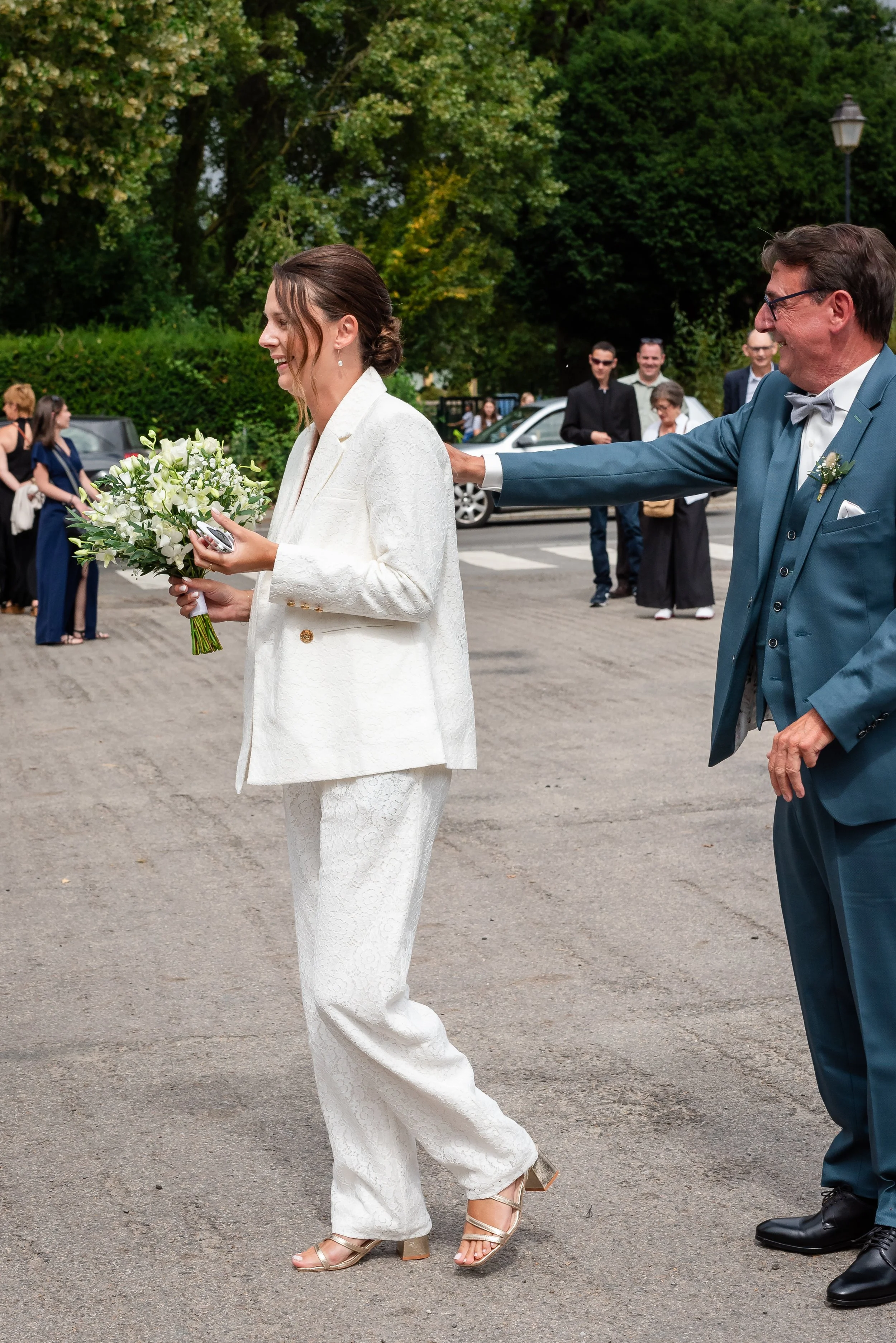 Une femme en costume blanc avec un bouquet de fleurs et un groupe de personnes en arrière-plan lors d'une célébration en plein air, probablement un mariage.