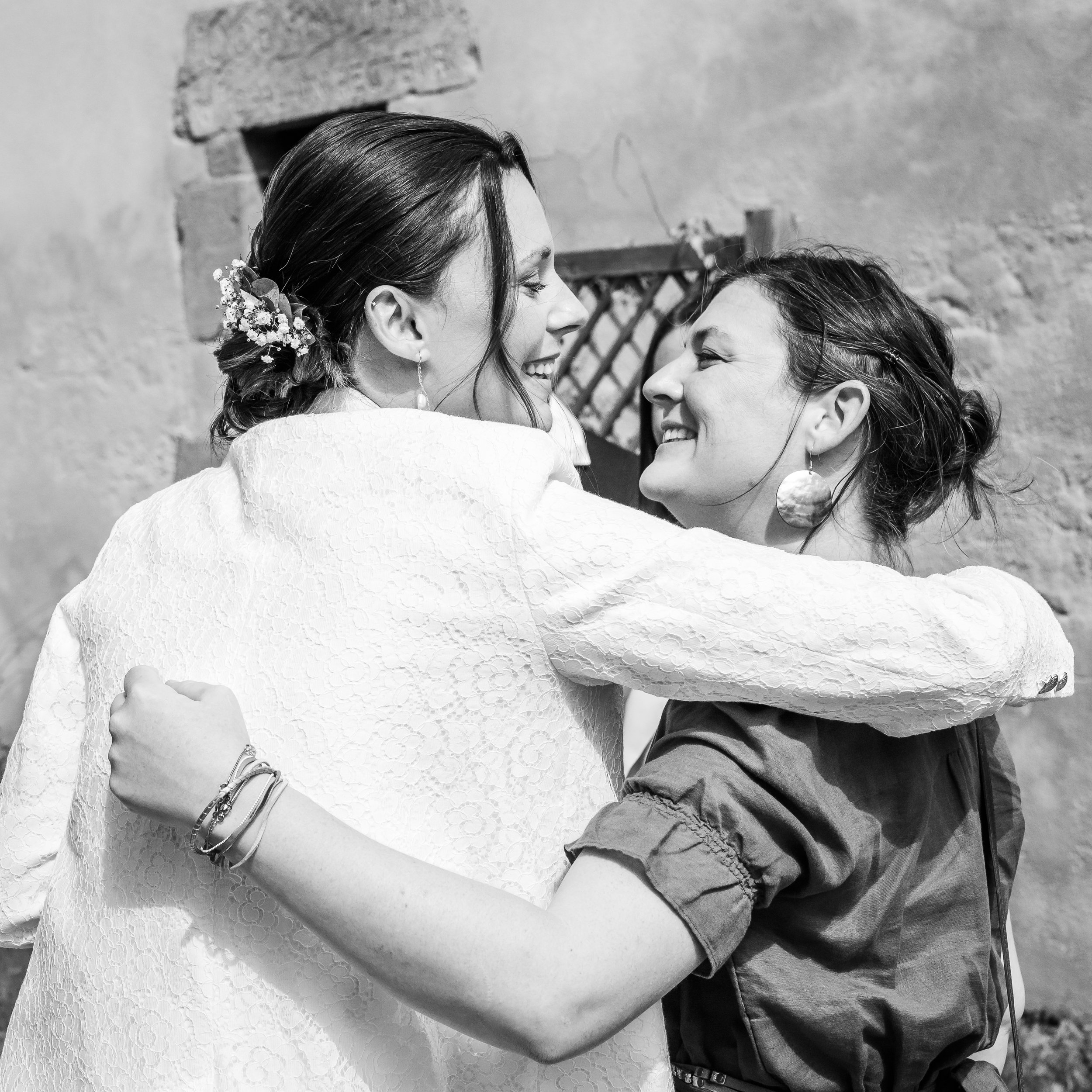 Deux femmes souriantes, l'une avec des fleurs dans les cheveux, en train de se faire un câlin, en noir et blanc.