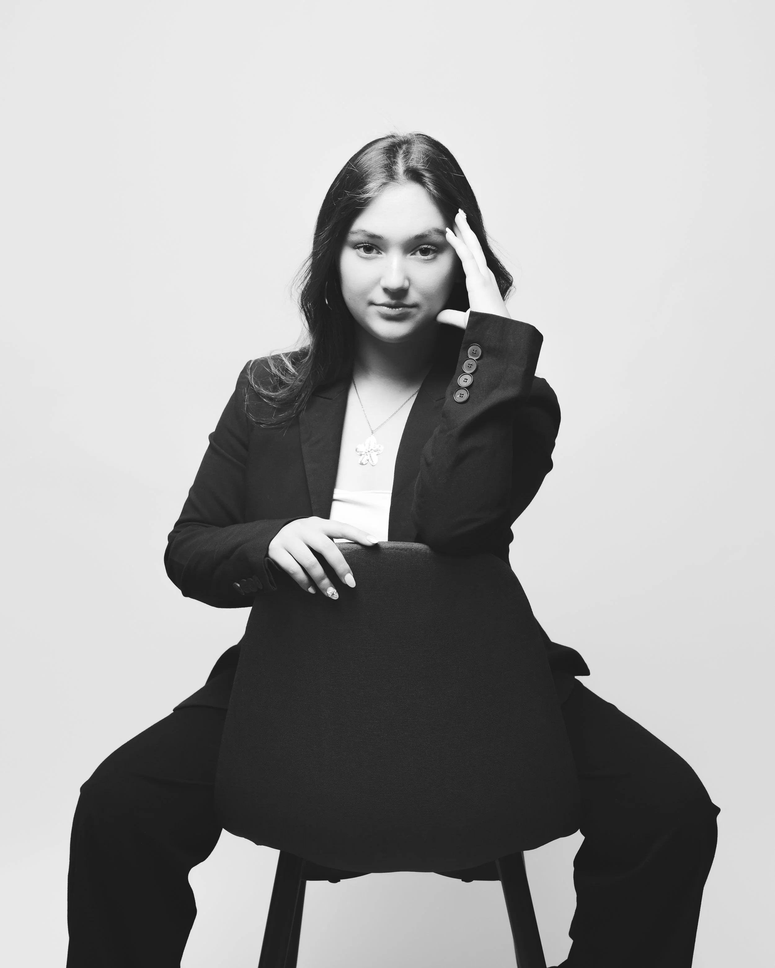 Une jeune femme en veste noire, assise sur une chaise, pose pour une photo en noir et blanc contre un fond blanc simple. Elle regarde directement l'appareil photo avec une expression confiante. Elle porte un collier en pendentif en forme de trèfle.