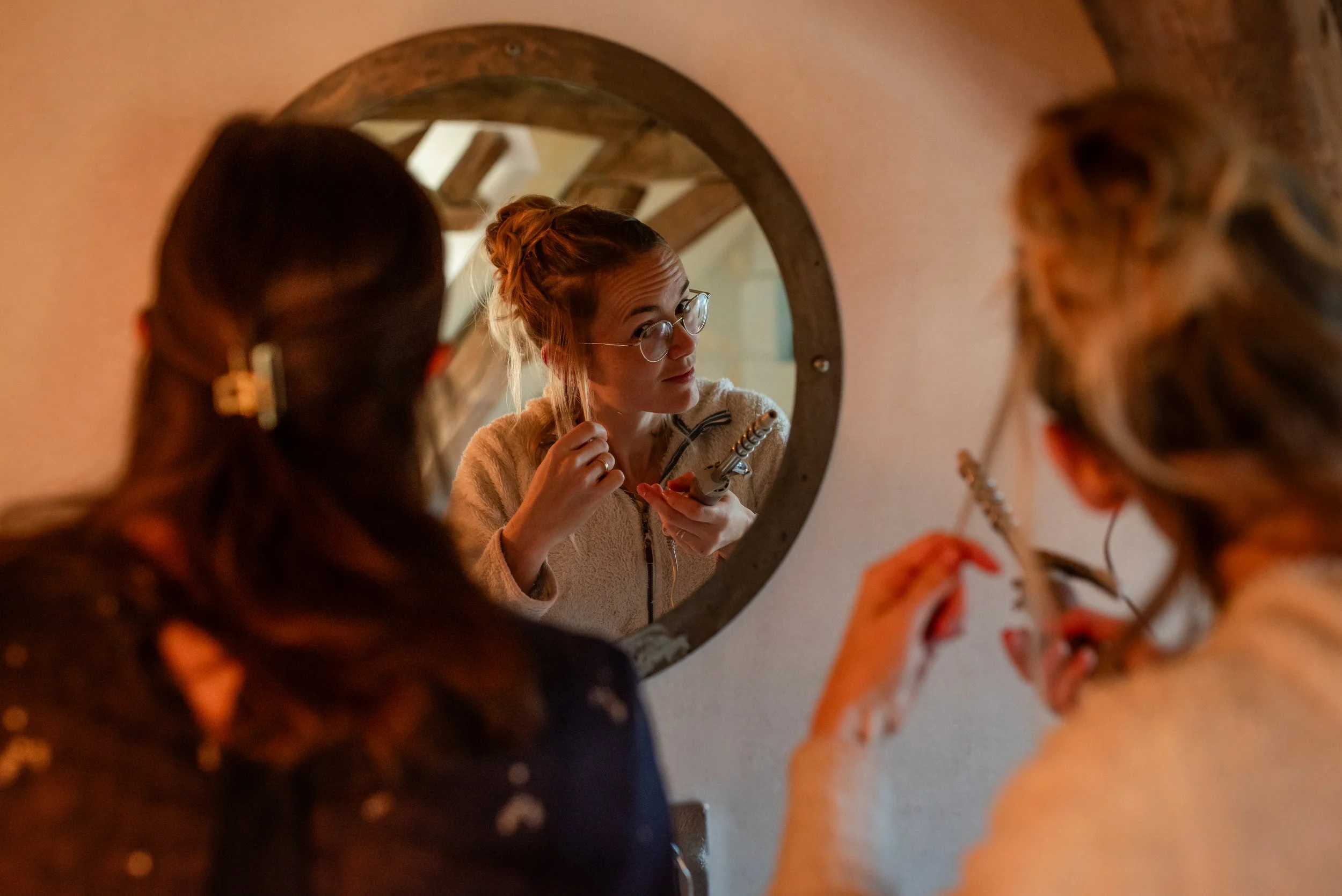 Une femme regarde dans un miroir rond, portant des lunettes et une veste beige, tandis que deux autres personnes, dont on voit la tête partiellement, sont présentes dans la pièce. La scène est capturée en intérieur avec un éclairage chaud.