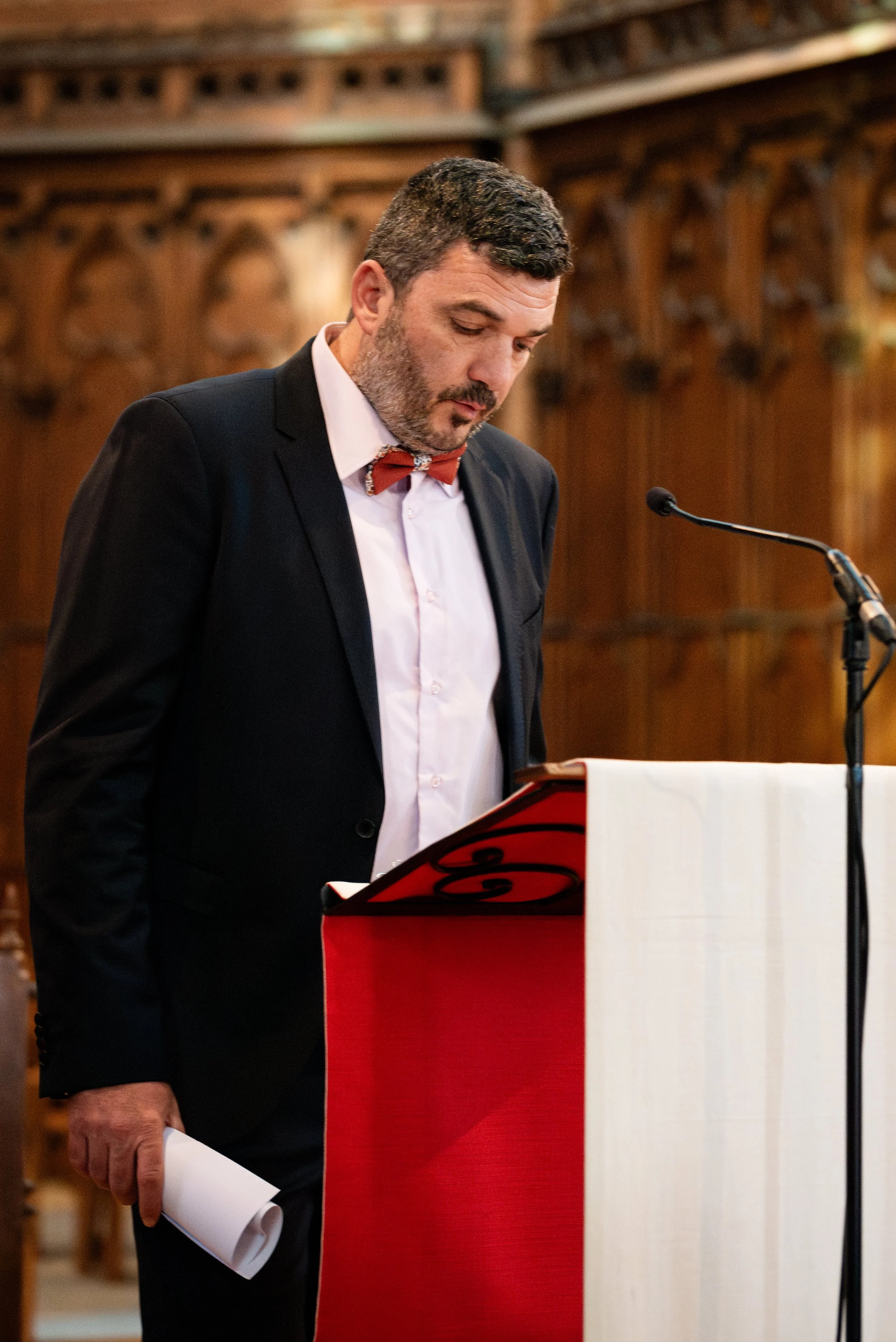 Homme en costume lisant un document lors d'une cérémonie ou d'un discours dans une salle ornée de bois.