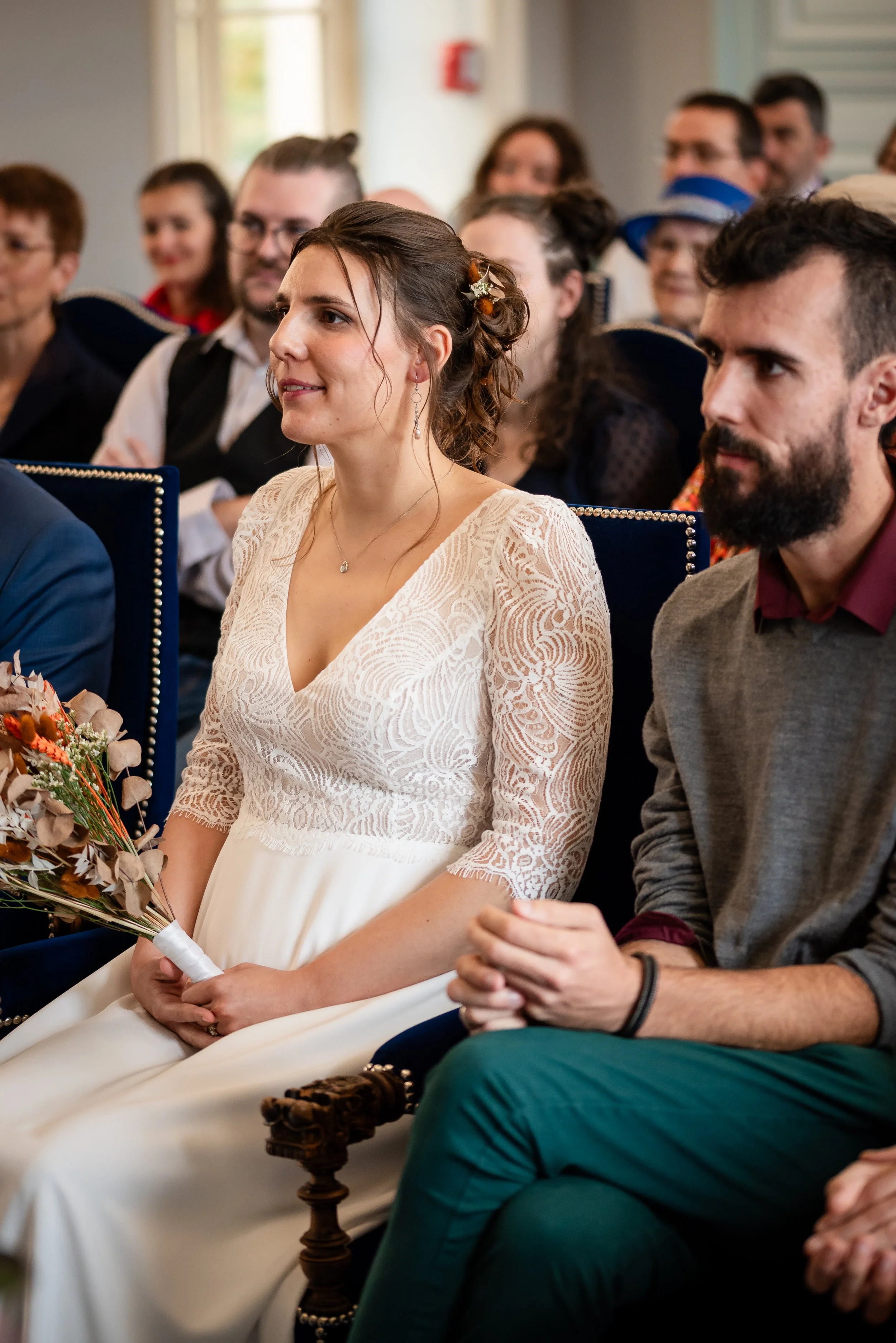 Une femme en robe blanche avec des détails en dentelle, assise lors d'une cérémonie, tenant un bouquet de fleurs aux couleurs automnales, entourée de personnes en tenue élégante.