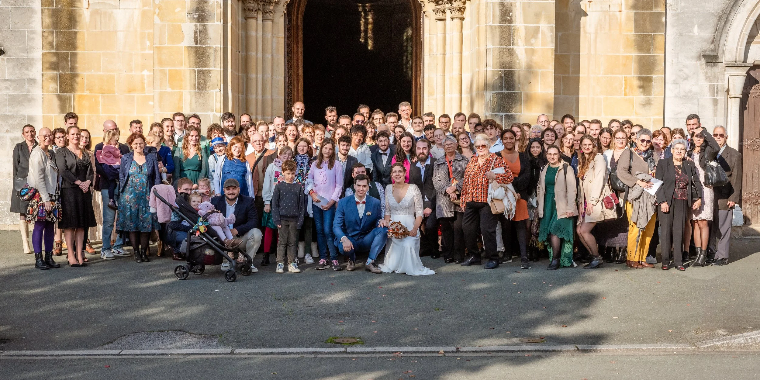 Gros groupe de personnes posant devant une façade de bâtiment en pierre, probablement pour une occasion spéciale comme un mariage.