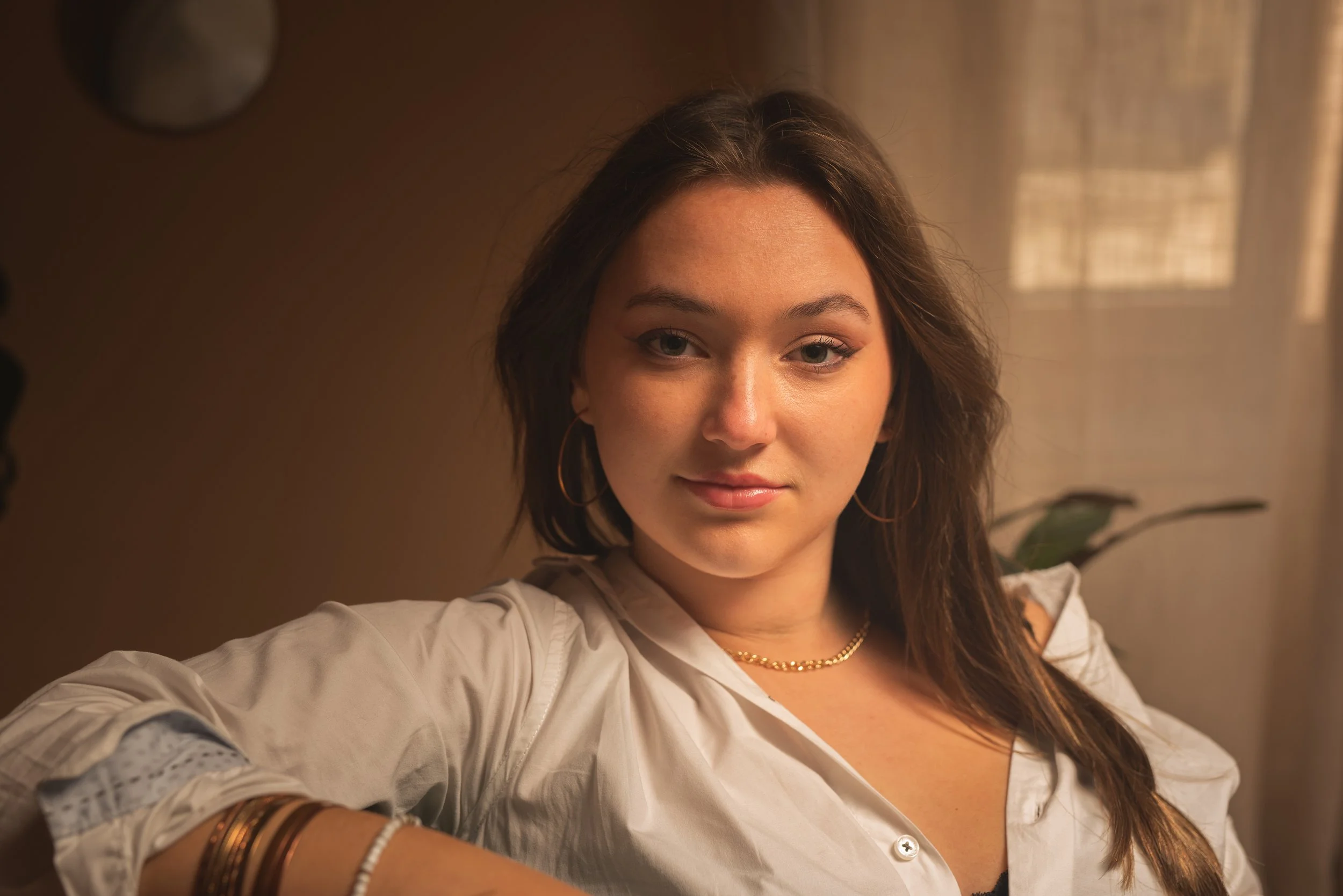 Jeune femme avec des cheveux longs et bruns, portant un chemisier blanc avec des manches roulées, des boucles d'oreilles en cercle, un collier en chaîne dorée, prenant un selfie dans une pièce éclairée par la lumière naturelle d'une fenêtre.