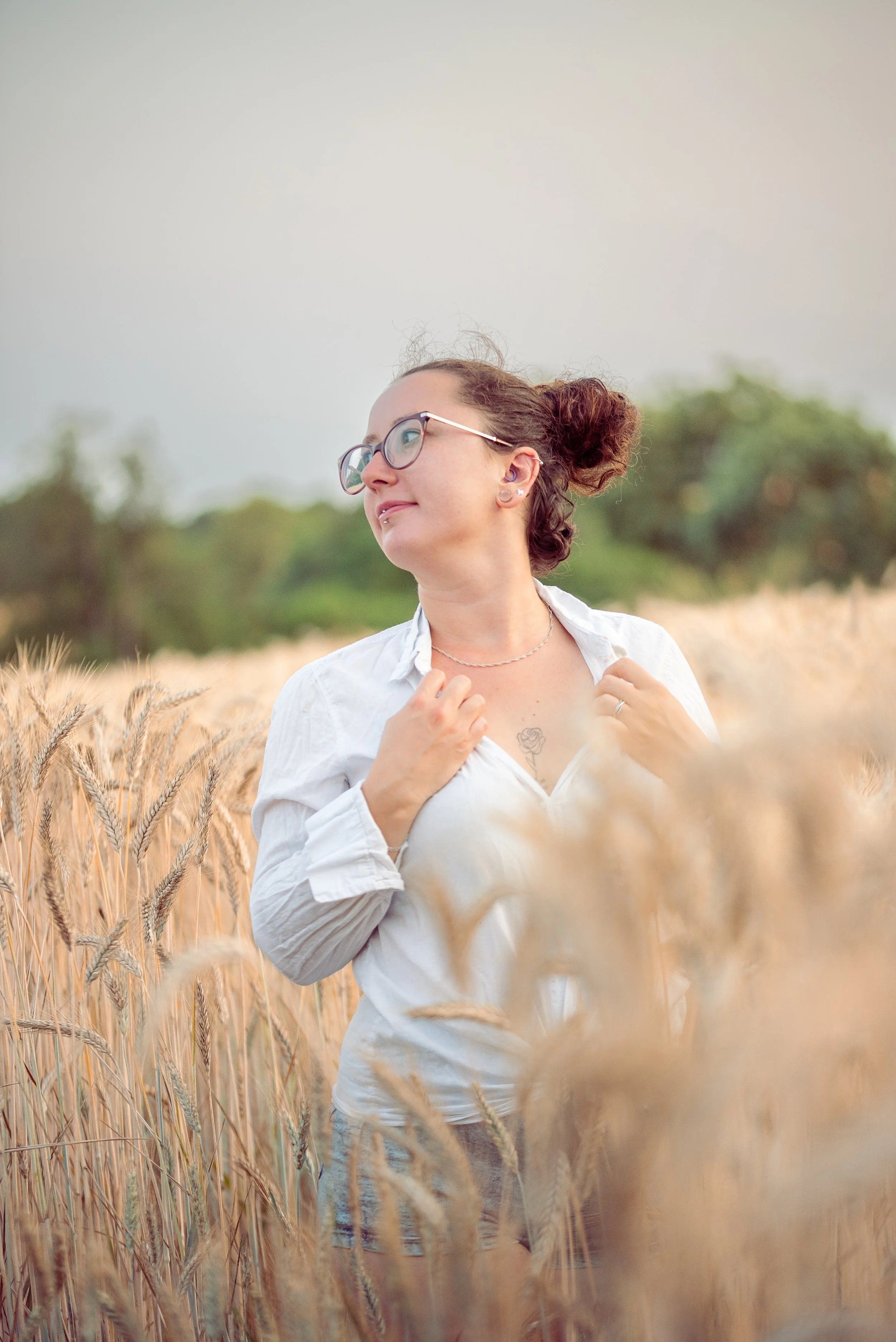 Jeune femme avec lunettes et piercing dans un champ de blé, portant une chemise blanche, regardant sur le côté, avec un fond de ciel gris et de végétation verte