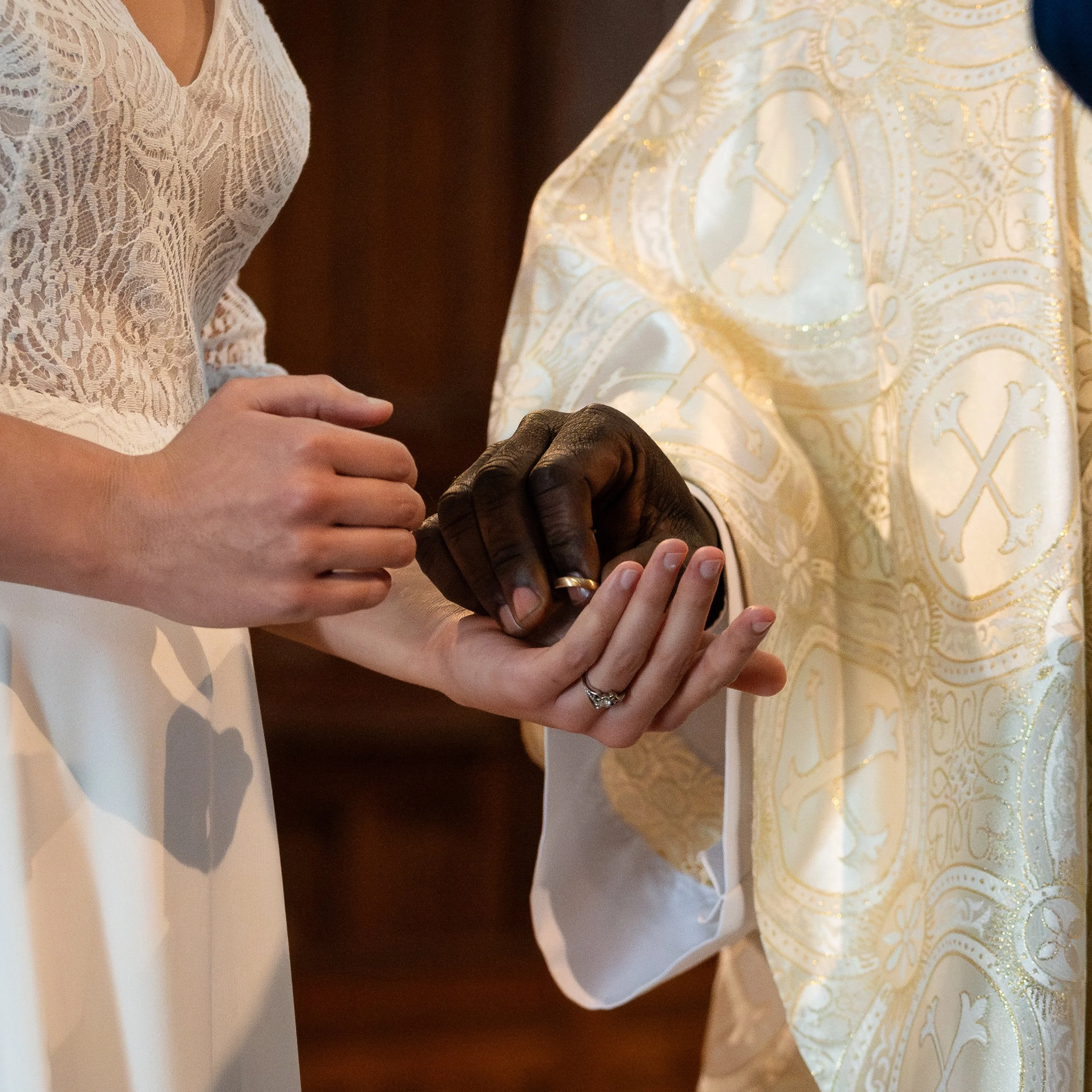 Deux personnes, dont une avec la peau foncée, échangent un anneau de mariage. La personne à peau foncée porte une bague et le bras d'une femme en robe de dentelle blanche. La scène a lieu près d'une table avec une nappe dorée à motifs.
