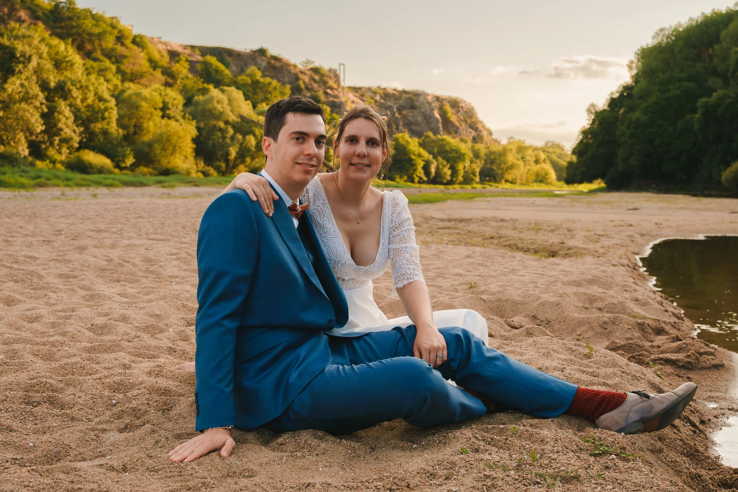 Un couple assis sur la plage, probablement lors de leur mariage, avec des montagnes verdoyantes en arrière-plan, au coucher du soleil.