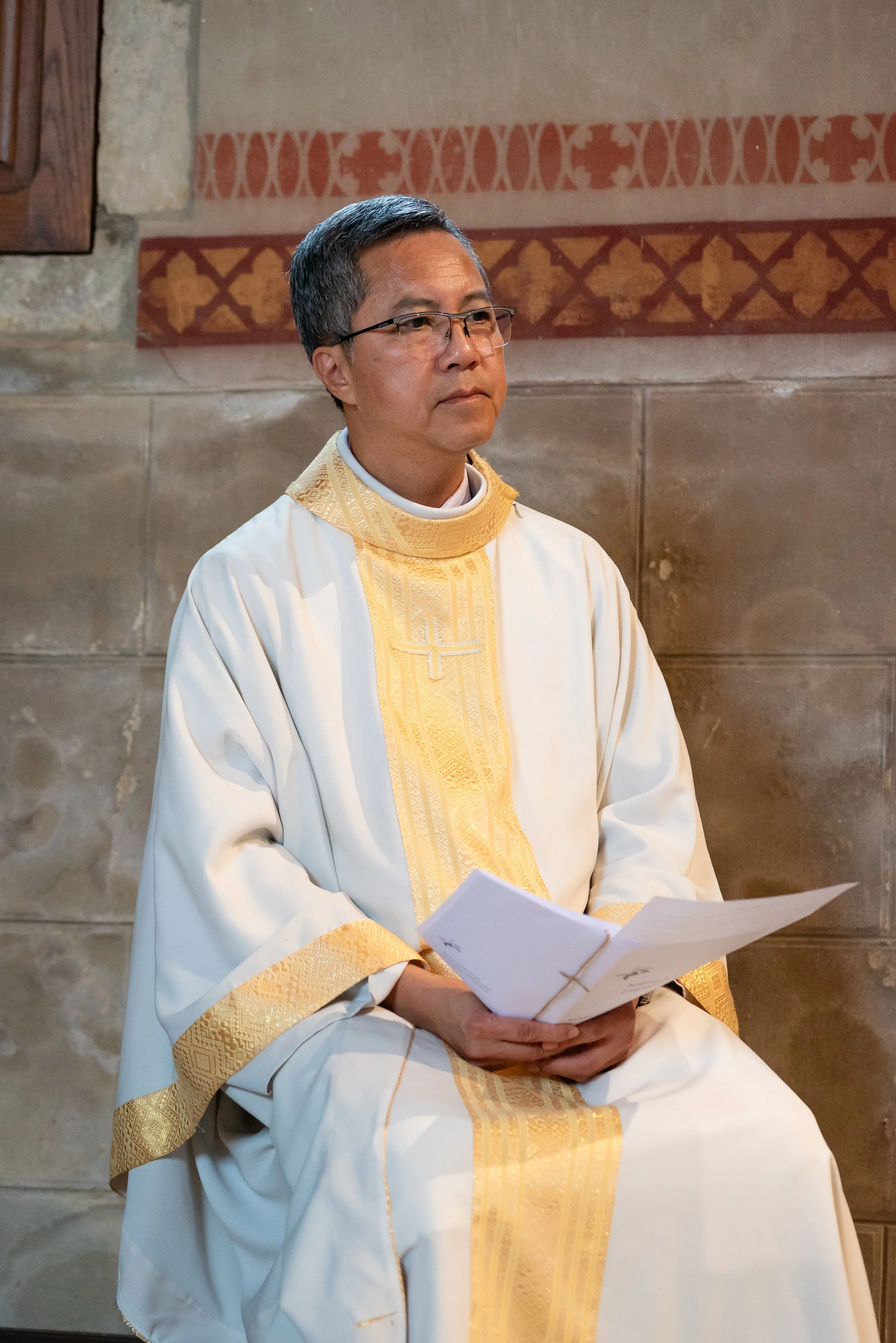 Un prêtre assis, portant des vêtements liturgiques blancs et dorés, tenant des papiers, dans une église en pierre avec fresques au mur.