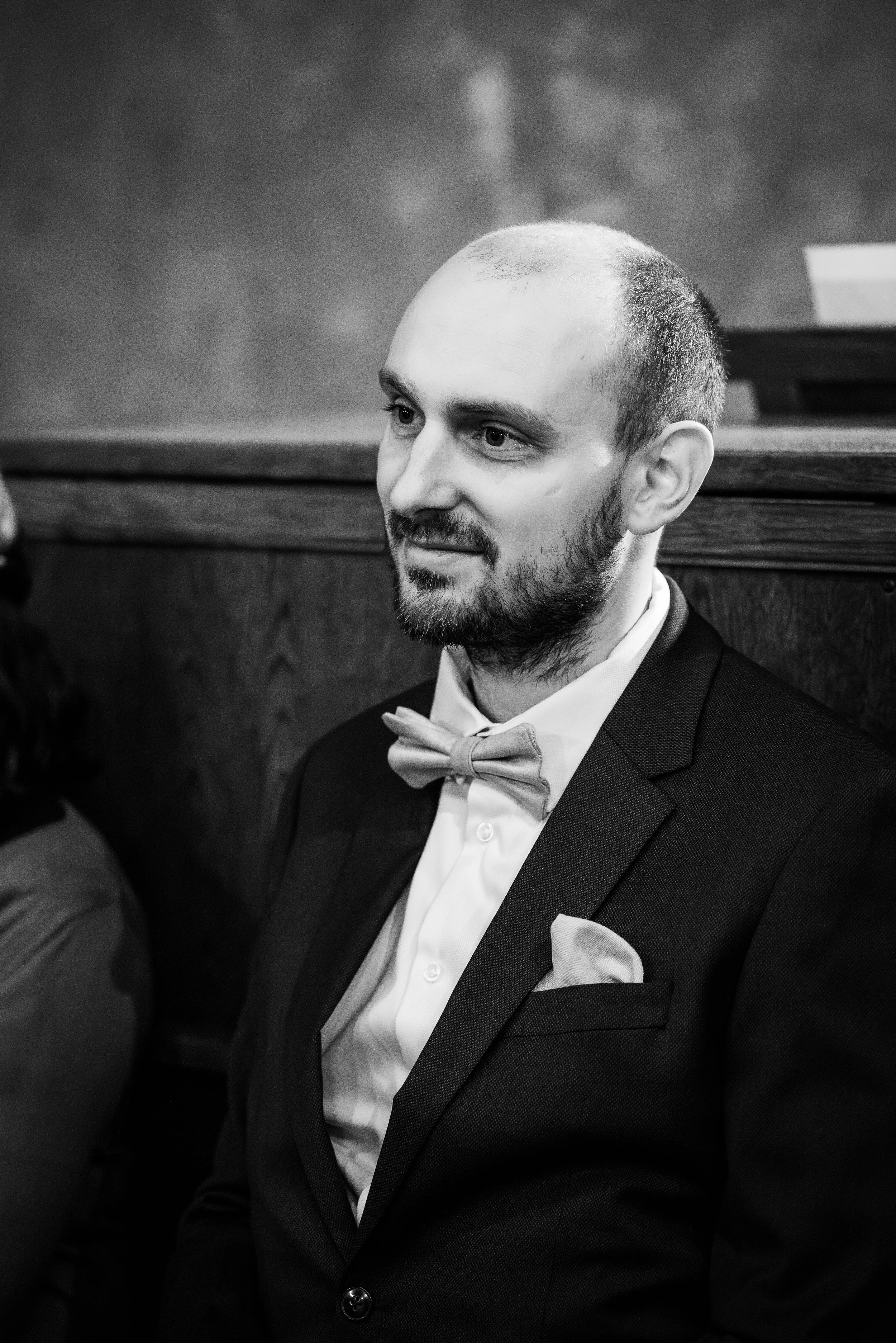 Un homme portant un costume noir avec un nœud papillon beige, assis dans une pièce avec un mur en bois derrière lui, regardant légèrement de côté, en noir et blanc.