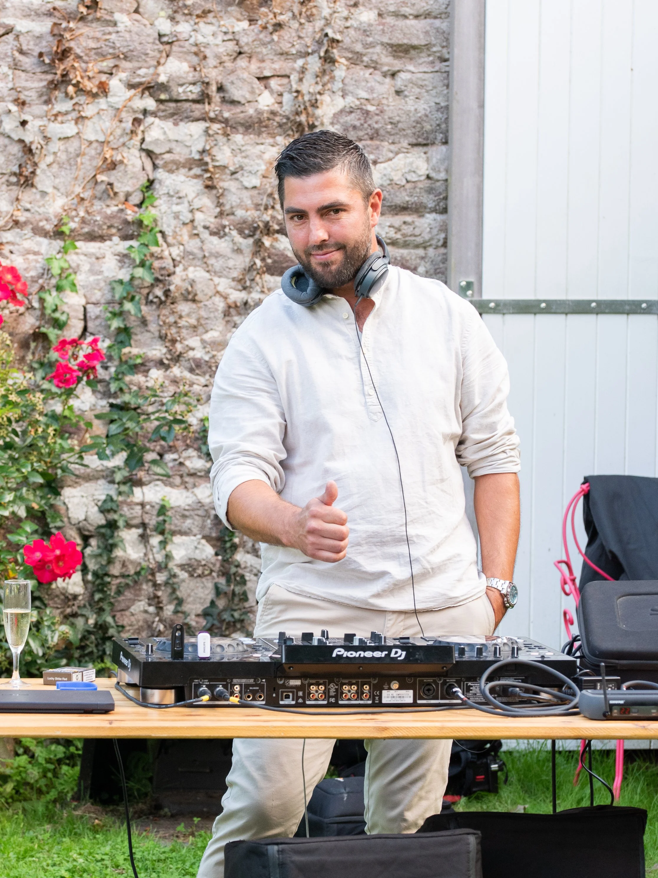 Un homme souriant, portant un casque autour du cou, se trouve derrière une table avec un DJ Pioneer DJ et du matériel audio, dans un jardin, donnant un pouce levé.