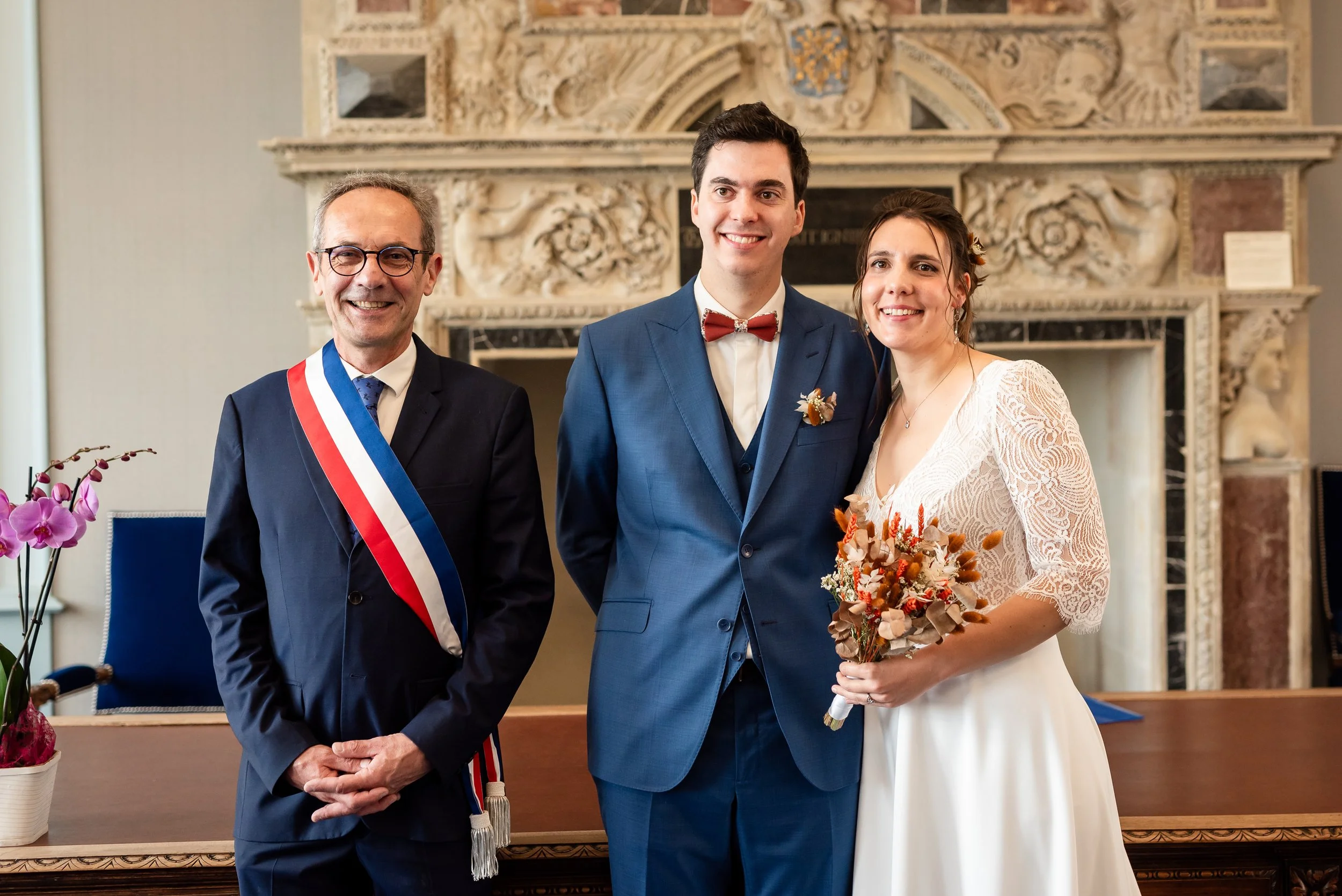 Un couple marié posant pour une photo au mariage, accompagnée d'une personne officielle portant une écharpe tricolore. Rapidements, on remarque une décoration intérieure sobre avec une cheminée en arrière-plan et une orchidée rose à gauche.