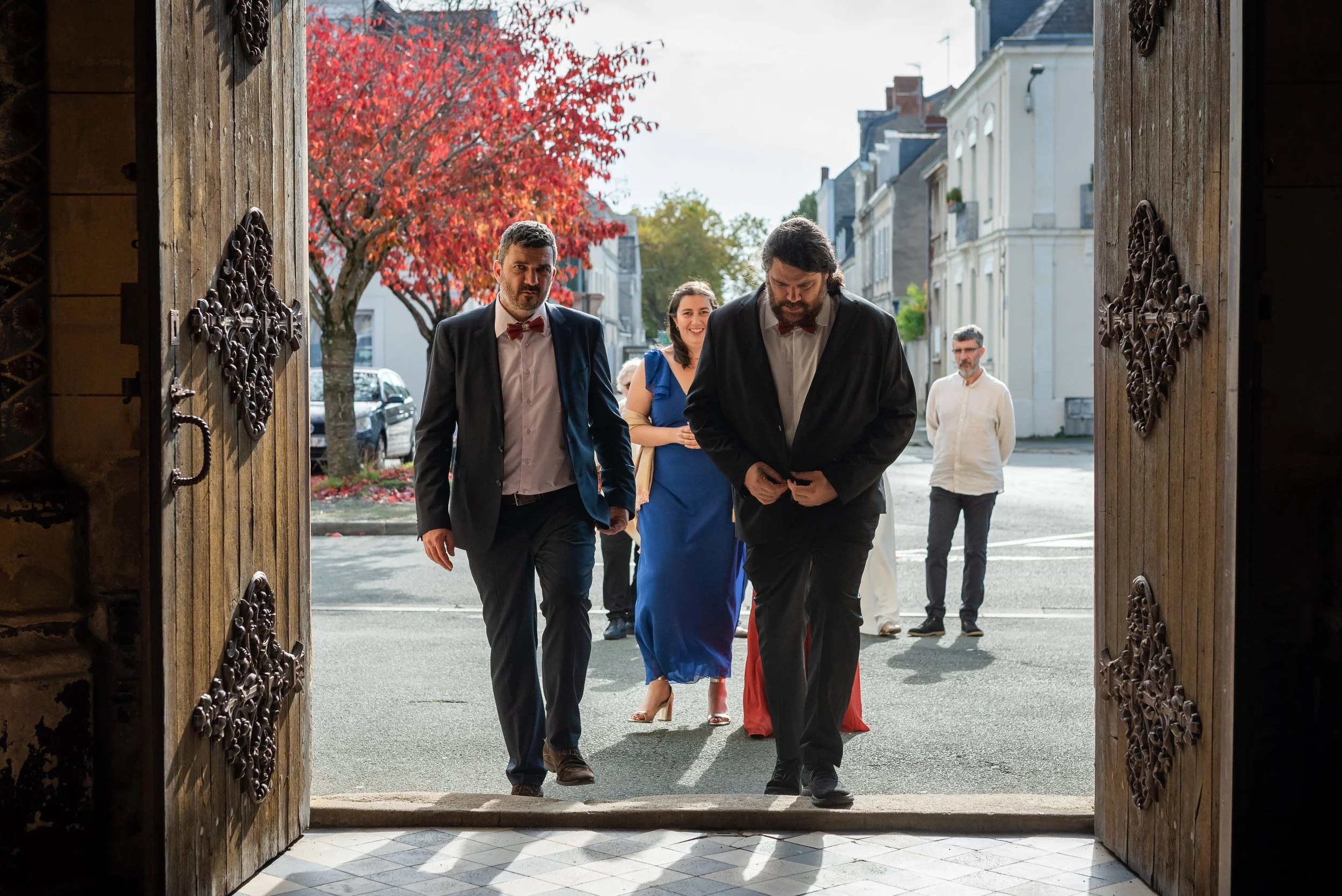 Groupe de personnes habillées de façon formelle sortant d'un bâtiment à l'entrée de la ville, avec des bâtiments résidentiels et un arbre aux feuilles rouges en arrière-plan.