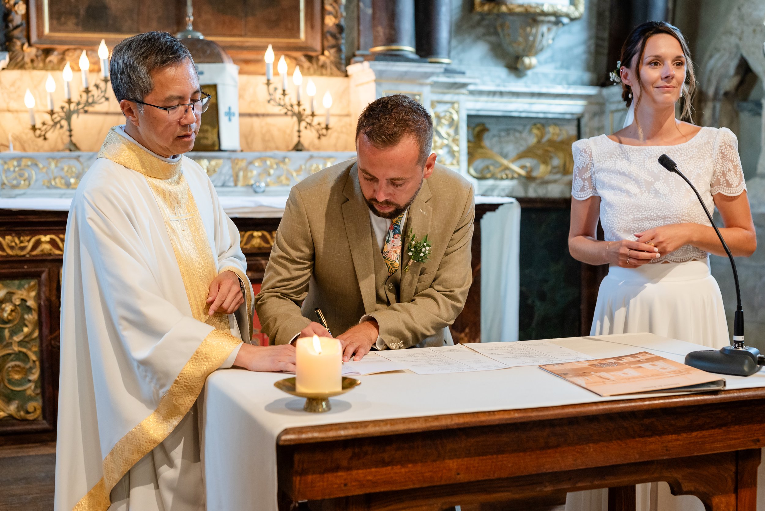 Un homme en costume beige signe un document lors d'une cérémonie religieuse dans une église ornée de chandeliers et de décorations dorées, avec un prêtre en robe blanche et une femme en robe blanche à côté.