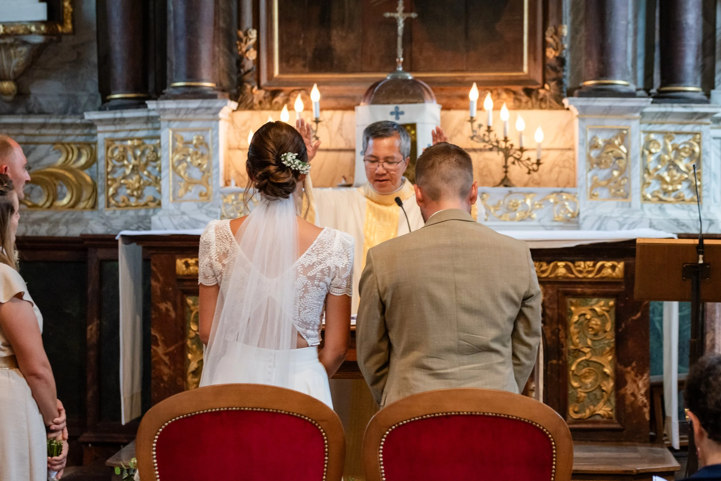 Un couple marié lors de leur cérémonie à l'autel dans une église décorée de chandeliers et de dorures, avec le prêtre au centre.