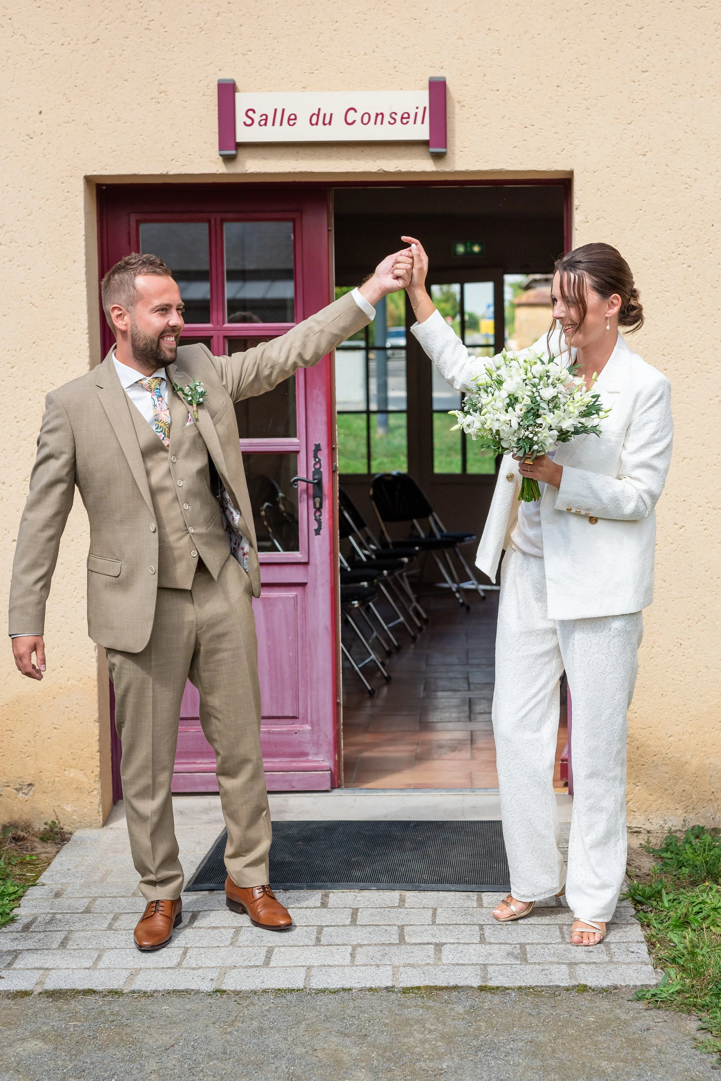Un couple de mariés, un homme en costume beige et une femme en tailleur blanc, s'apprêtent à entrer dans une salle de conseil, en se tenant la main et en souriant, devant une porte violette.