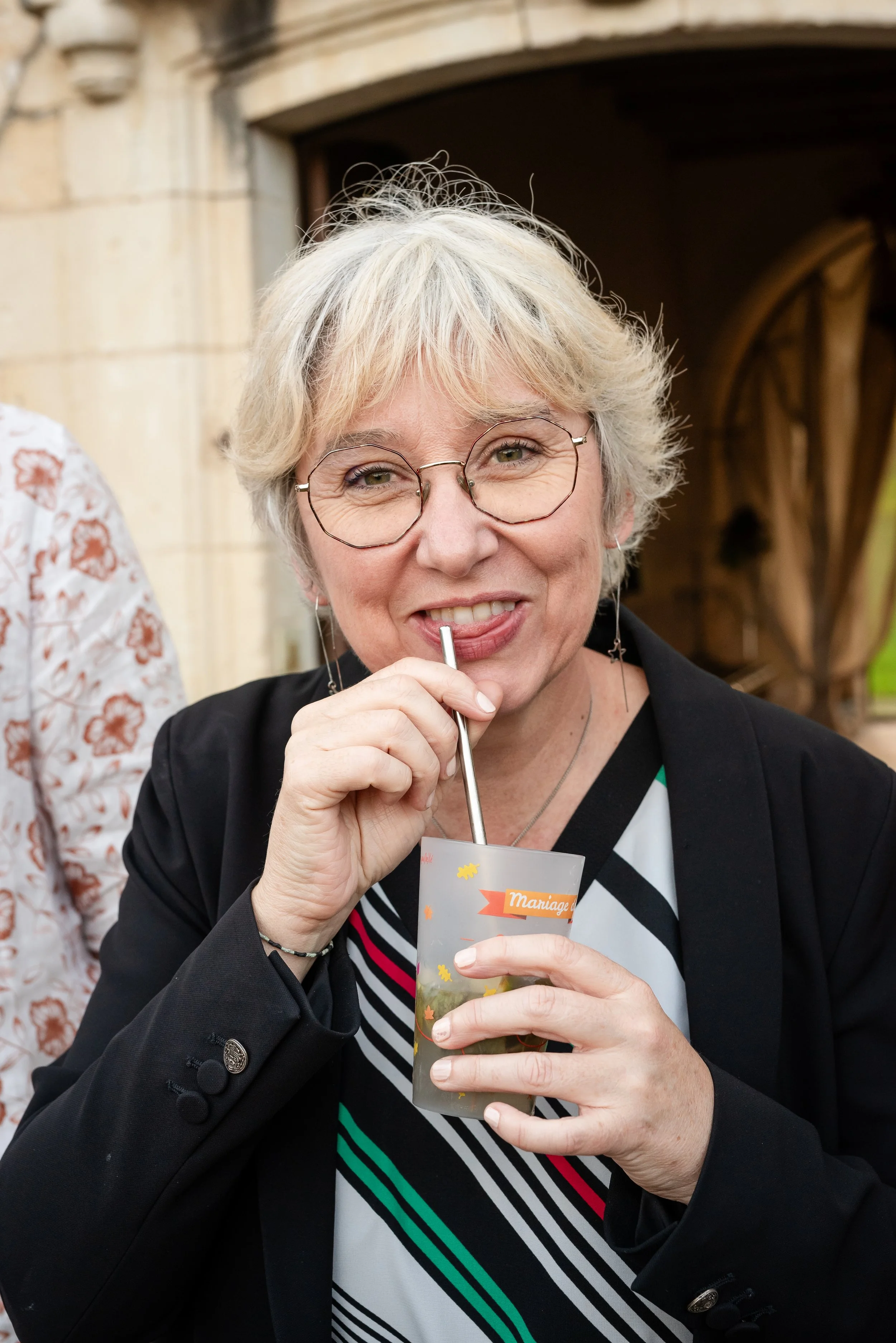 Femme à cheveux gris, portant des lunettes rondes, souriante, tenant un verre de boisson avec une paille, en extérieur avec un bâtiment en pierre en arrière-plan.