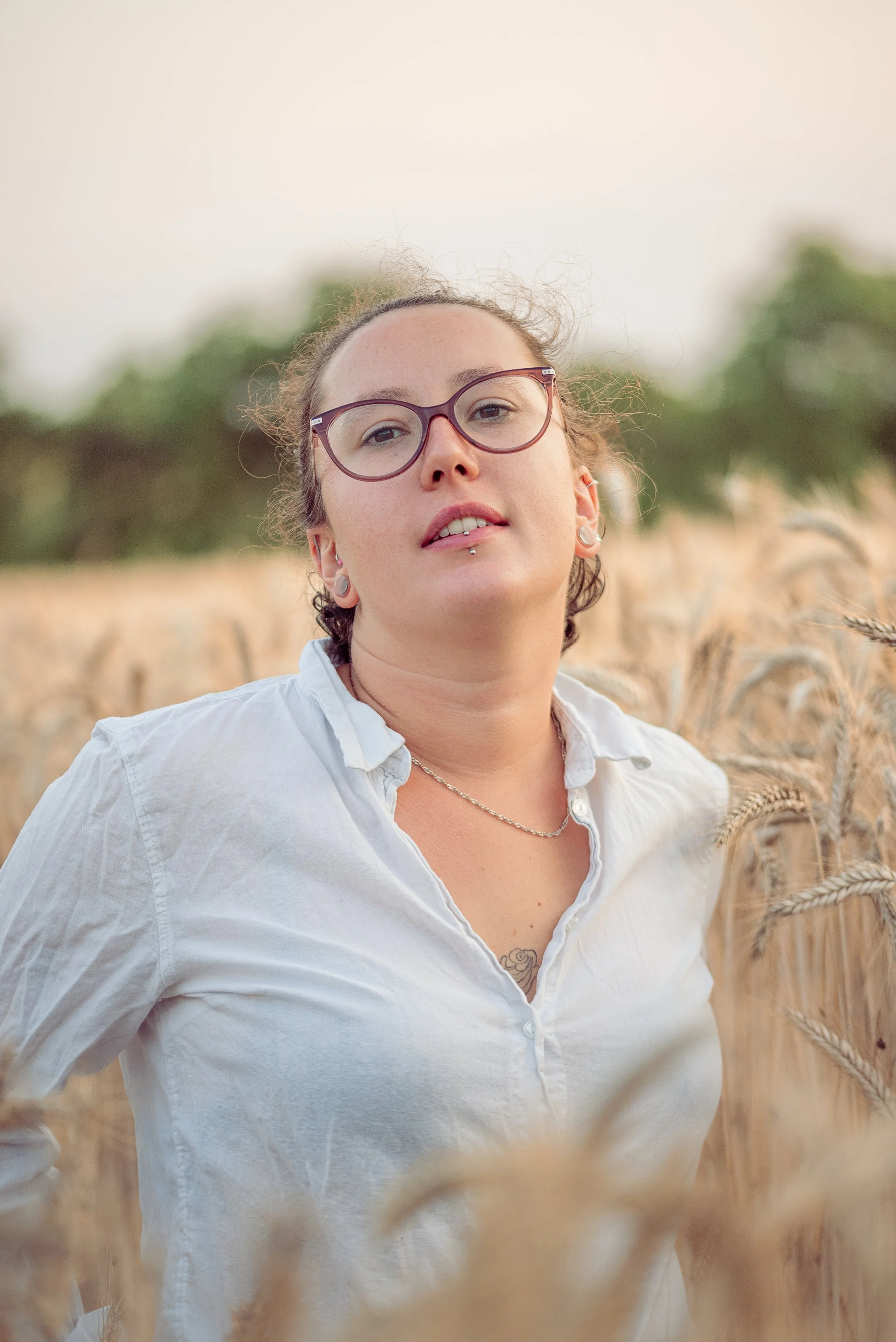 Une femme avec des lunettes, des boucles d'oreilles et une chaîne, portant une chemise blanche, se tient dans un champ de blé.