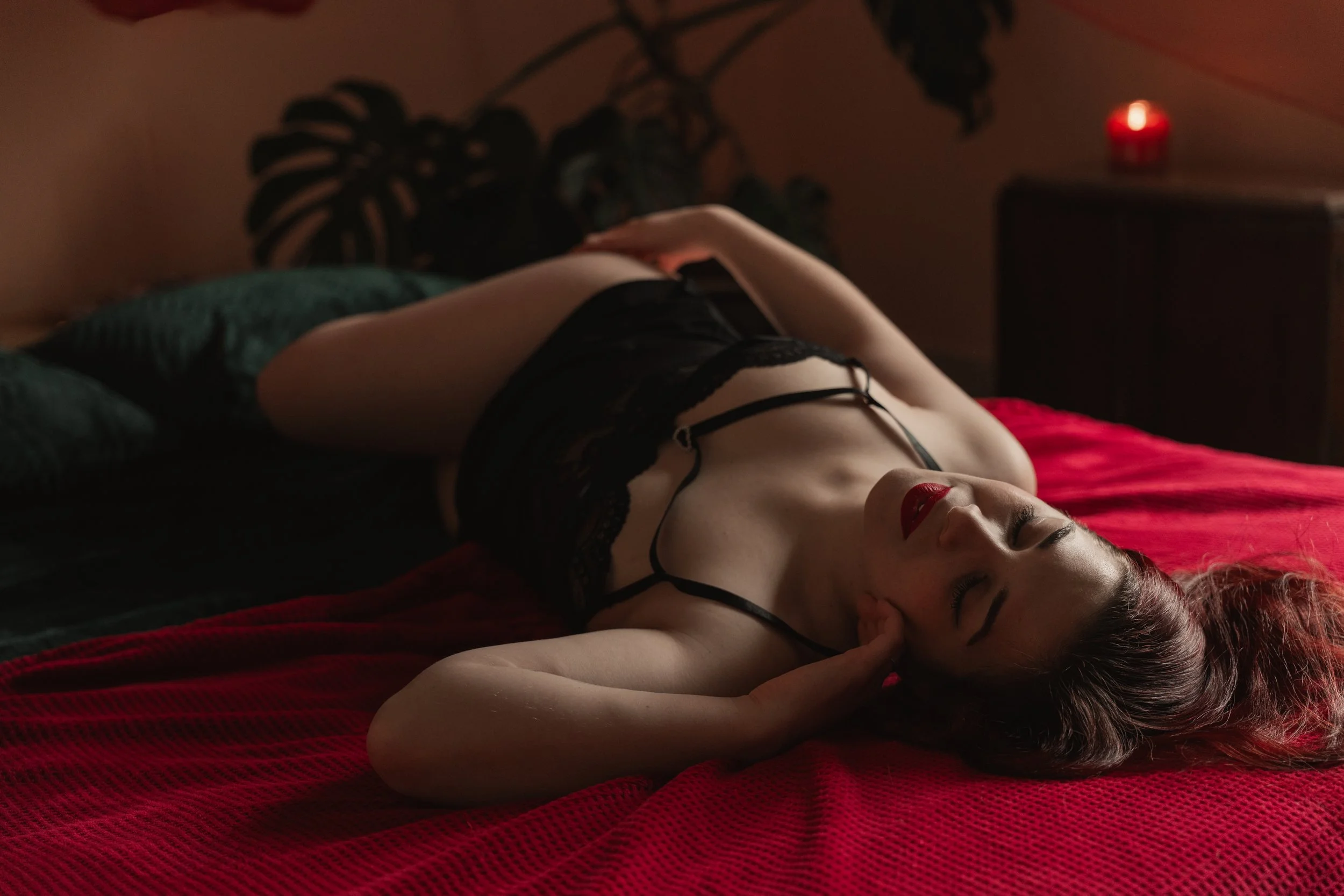 Une femme allongée sur un lit rouge, portant de la lingerie noire avec des bretelles. Elle a les yeux fermés et semble détendue, avec une main près de sa tête. La pièce est tamisée, avec un fond flou et une veilleuse rouge sur une table.